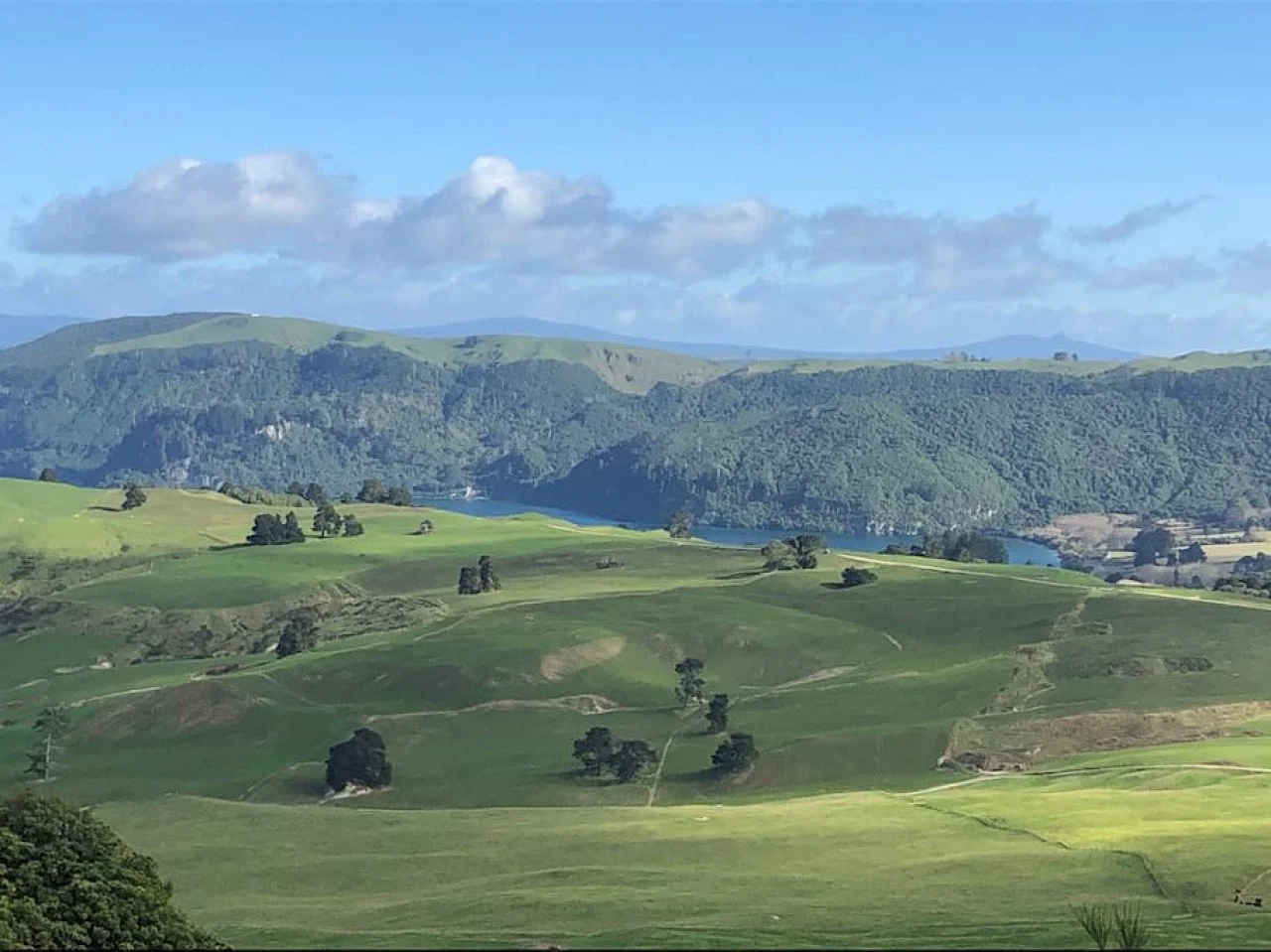 Scenic landscape with rolling green hills, trees, a river, forested mountains in the background, and a cloudy blue sky.