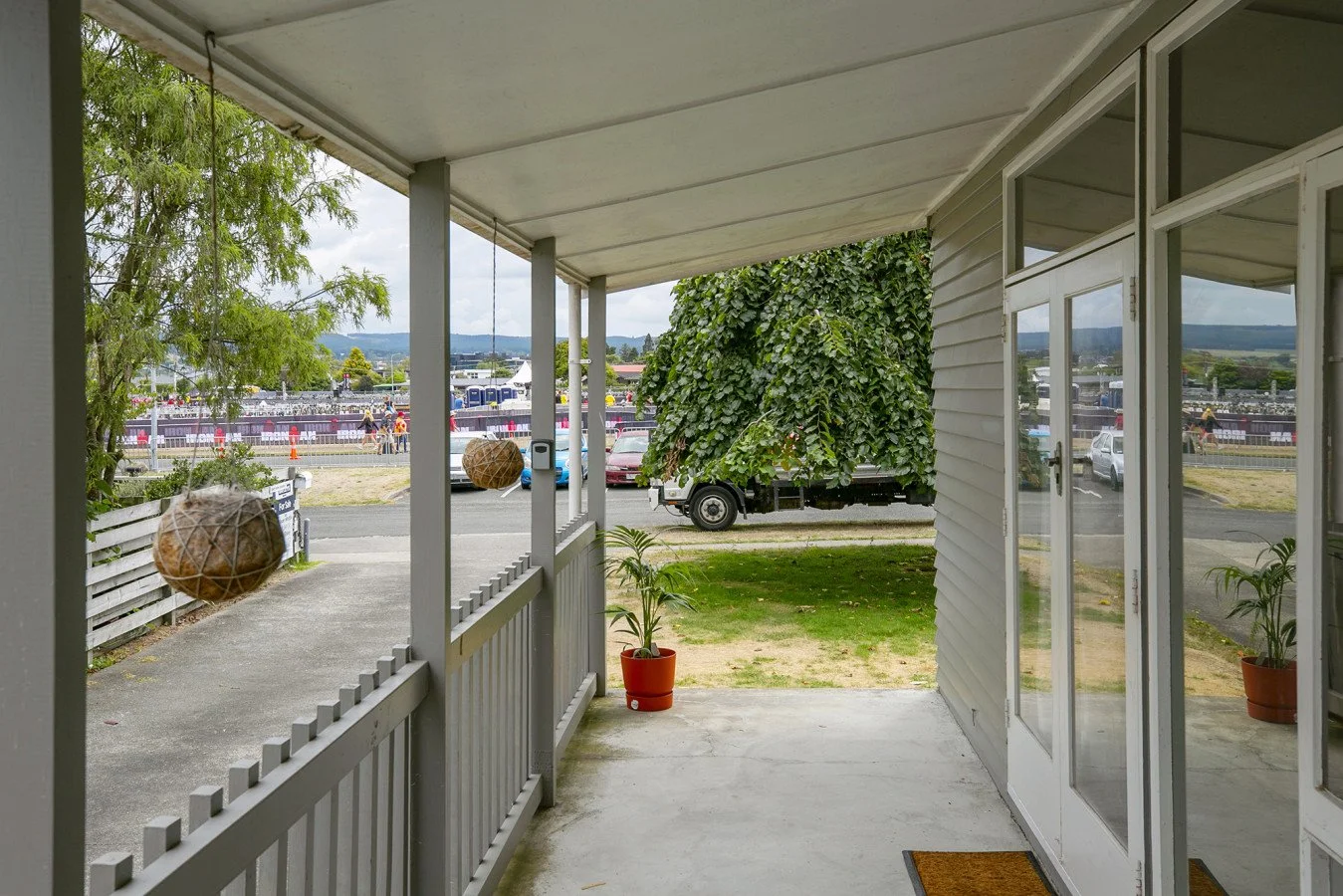 View from a porch with potted plants, looking out onto a street with parked cars, trees, and a large event with tents and people in the background.
