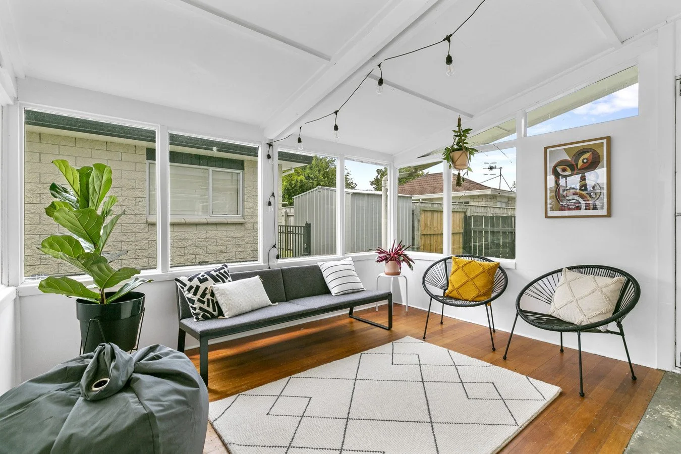 Sunroom with white walls and ceiling, hardwood floor, large windows, and minimal decorated seating area including a gray sofa with striped and patterned pillows, two black wire chairs with cream and yellow cushions, a small white table with a plant, 
