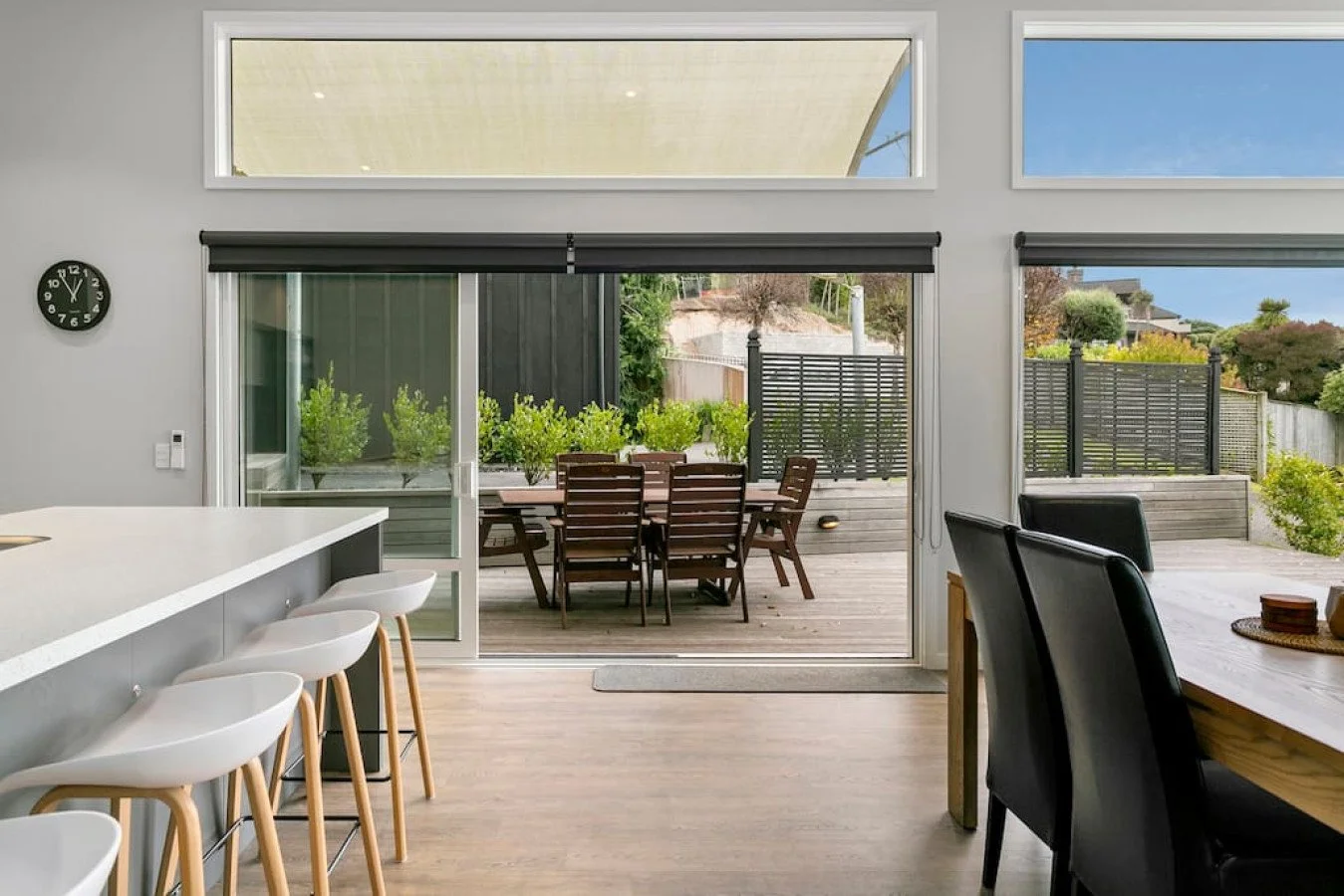Interior view of a modern dining area with a sliding glass door opening to an outdoor wooden deck with a dining table and chairs, surrounded by greenery and houses in the background.
