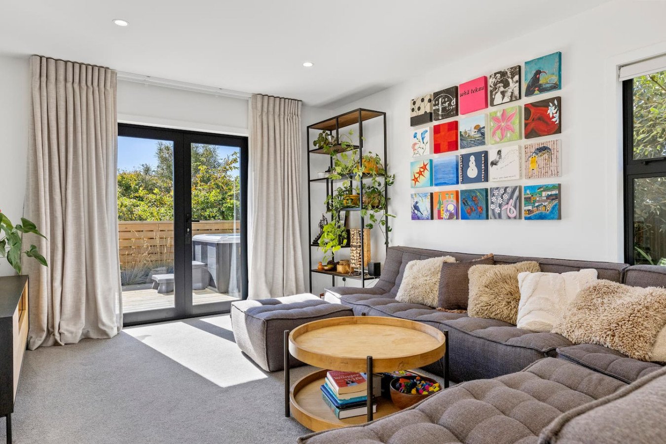 Living room with large sectional gray sofa, fluffy pillows, round wooden coffee table with books, bookshelf with plants, and a grid of colorful artwork on white walls. Glass door opens to patio with outdoor furniture and greenery.