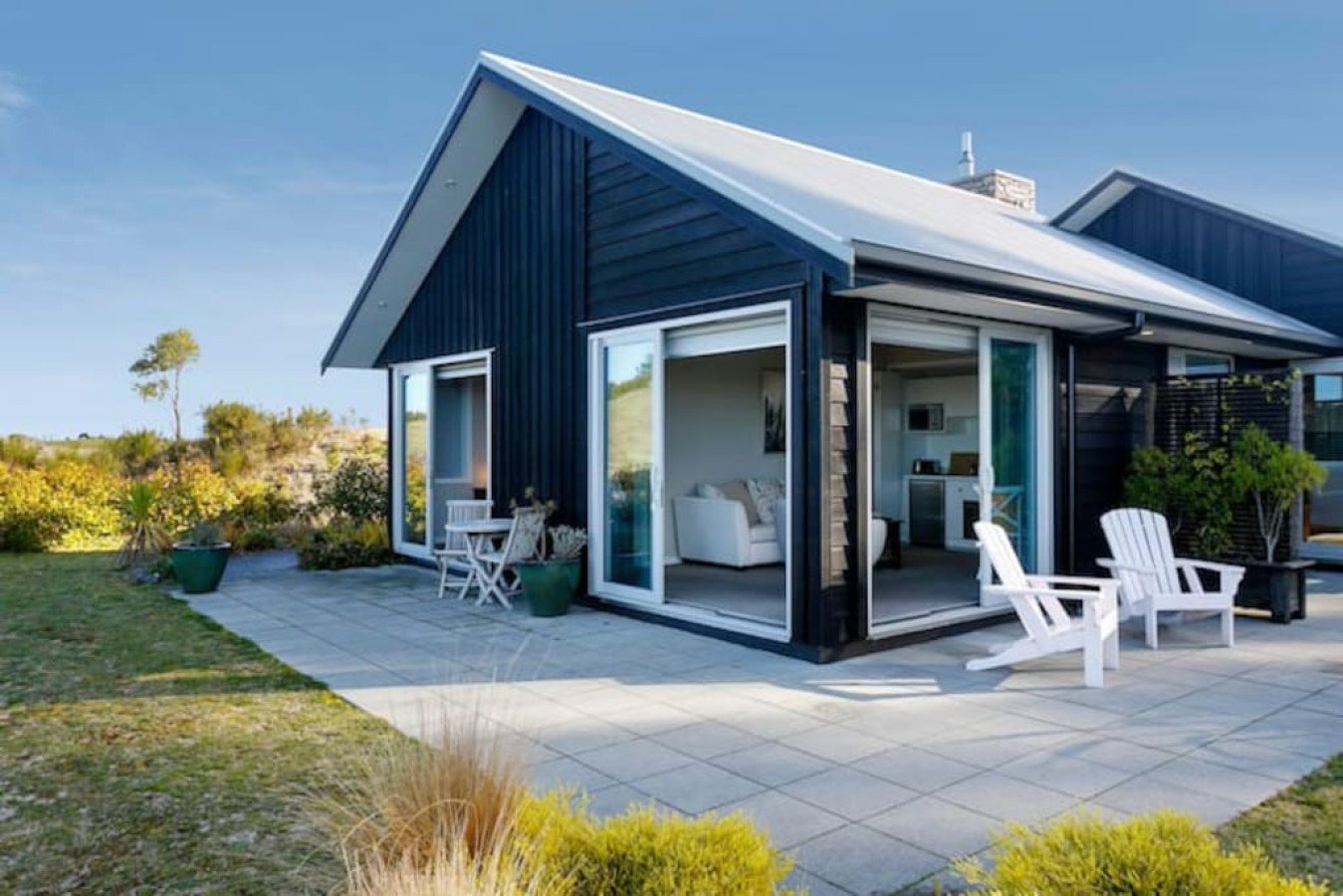 Modern black house with large sliding glass doors opening to a patio with white chairs, surrounded by greenery and a clear blue sky.