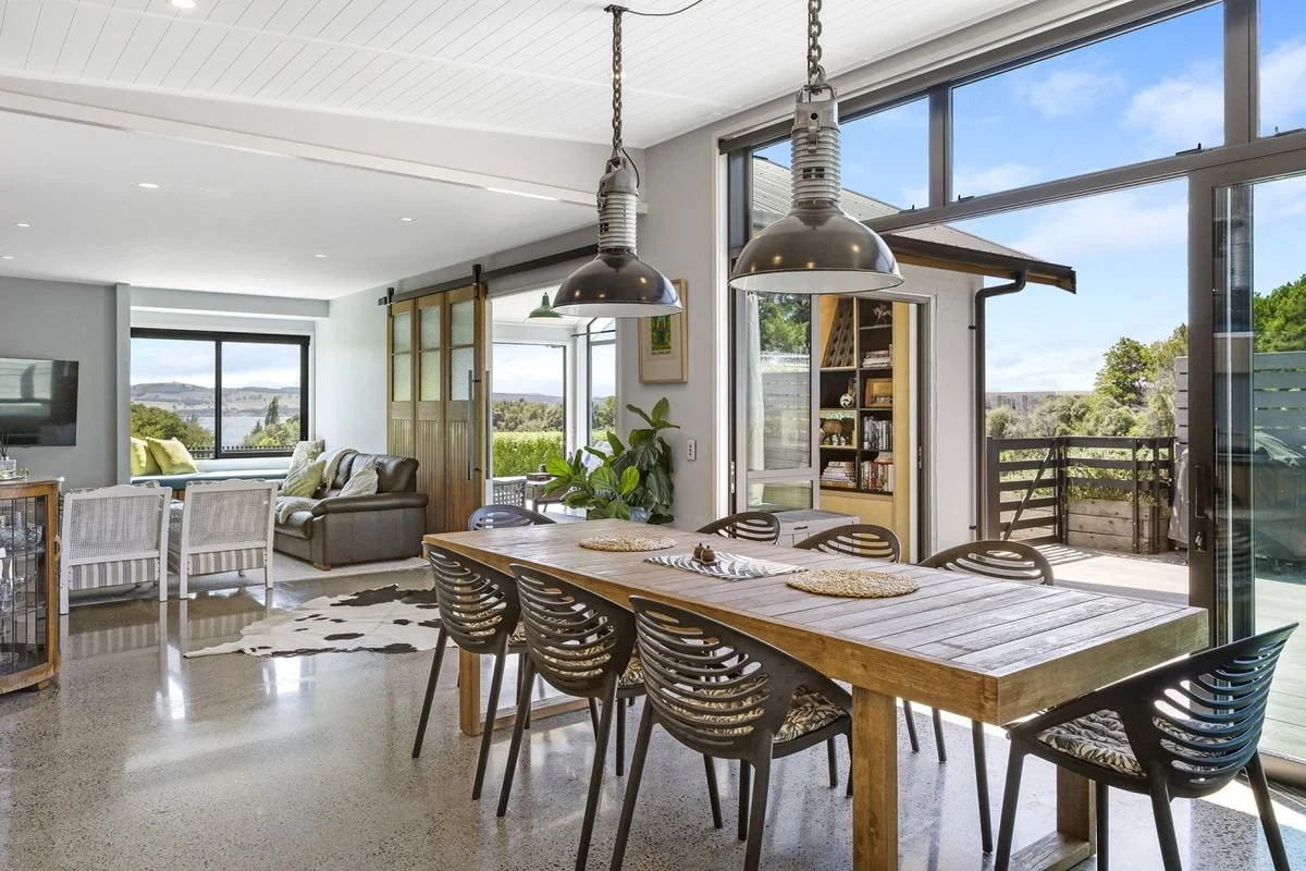 Open-concept living and dining area with large windows showing a view of greenery and water, featuring a wooden dining table with black chairs and a cozy living space with sofas and a TV.