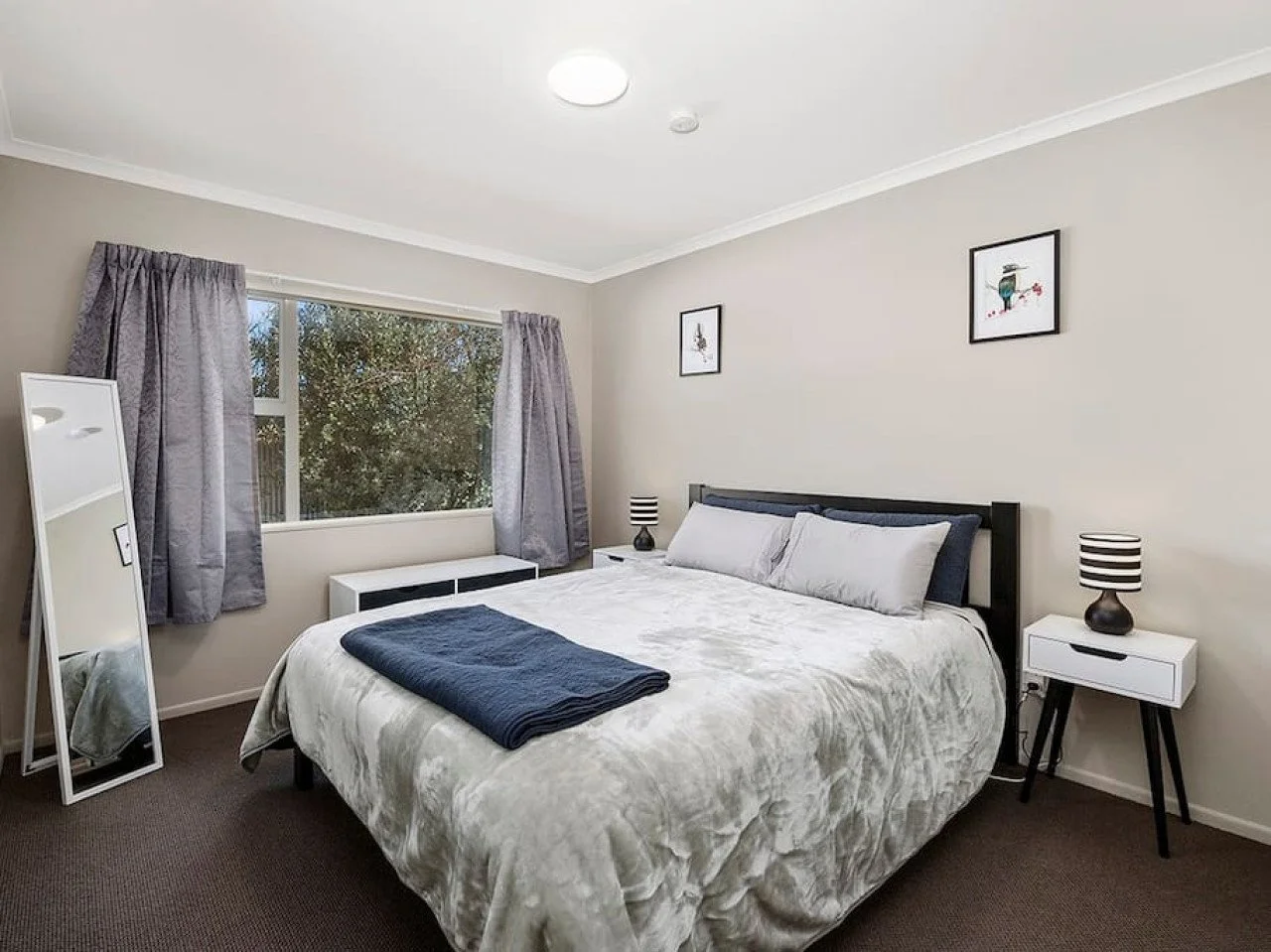 A neatly made bed with white bedding and gray pillows in a bedroom with beige walls, a window with gray curtains, two white nightstands with black lamps, a white full-length mirror, and framed artwork on the wall.