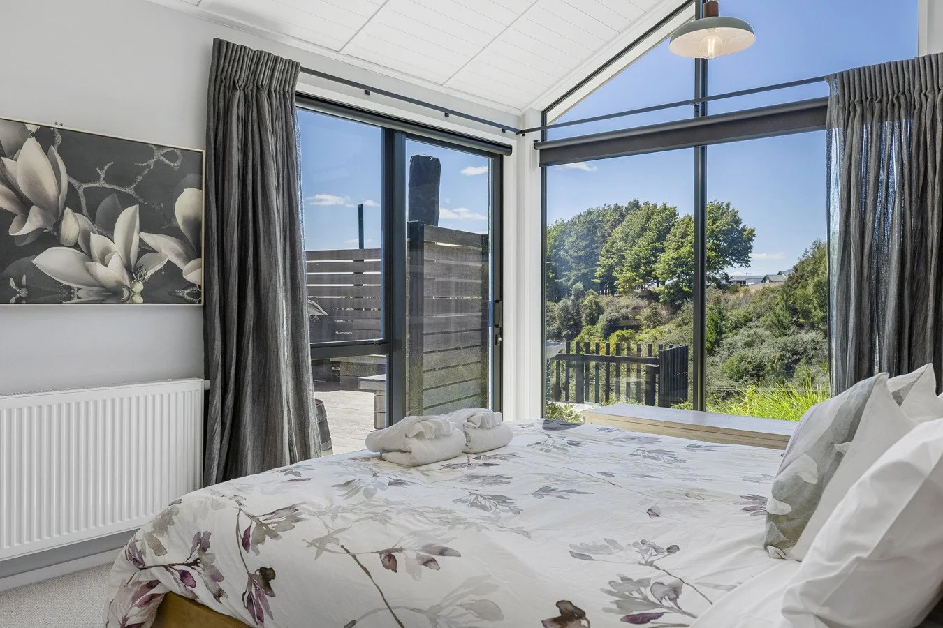 Bright bedroom with large windows showing a scenic outdoor view, bed with floral bedding, and towels folded on top.