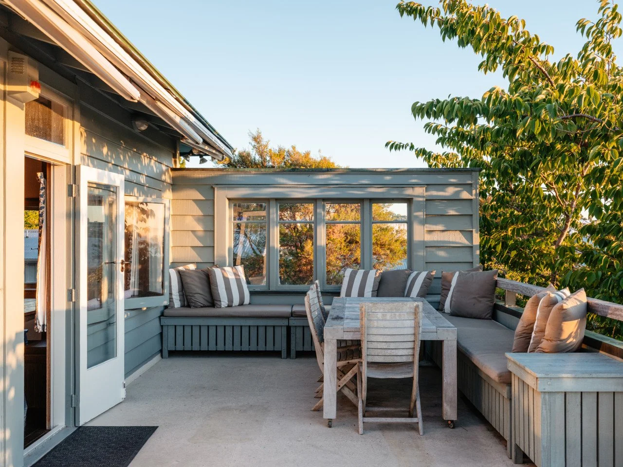 Outdoor patio with built-in bench seating, cushions, a wooden table, and chairs, surrounded by trees with sunlight casting shadows.