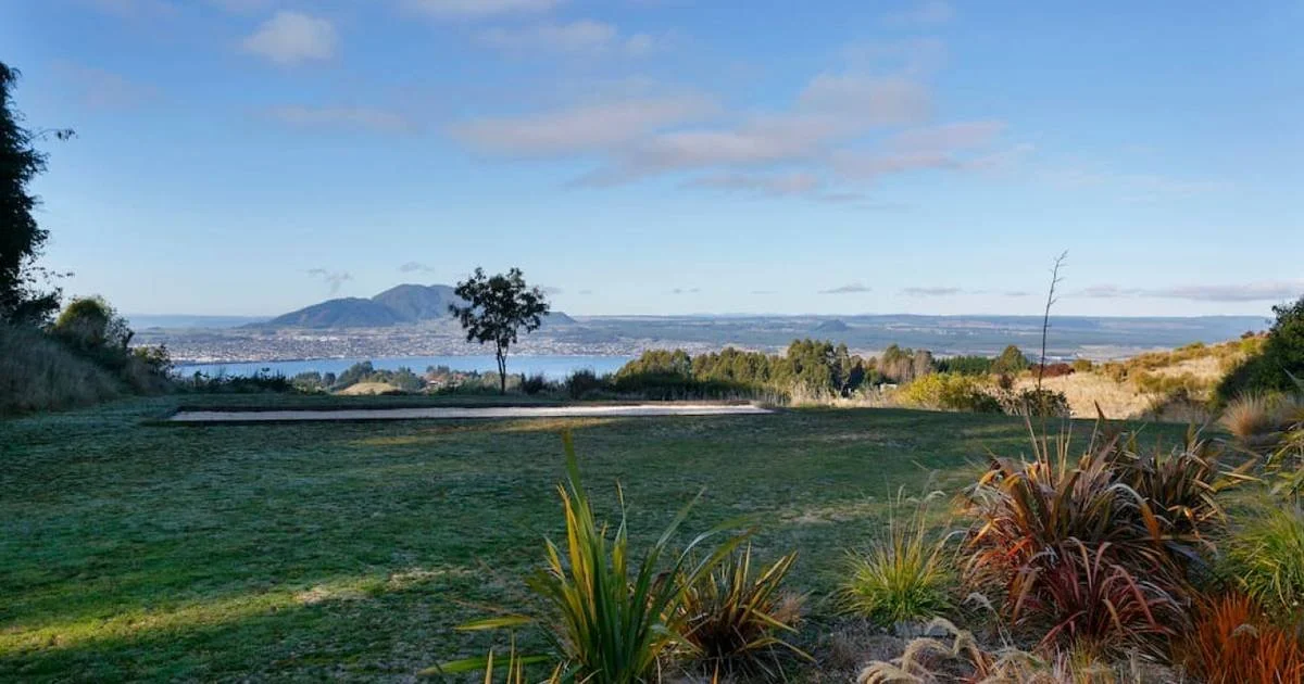 A scenic landscape with a grassy area in the foreground, a few trees, and a mountain in the background. There is a body of water visible to the left of the mountain, and the sky is partly cloudy.