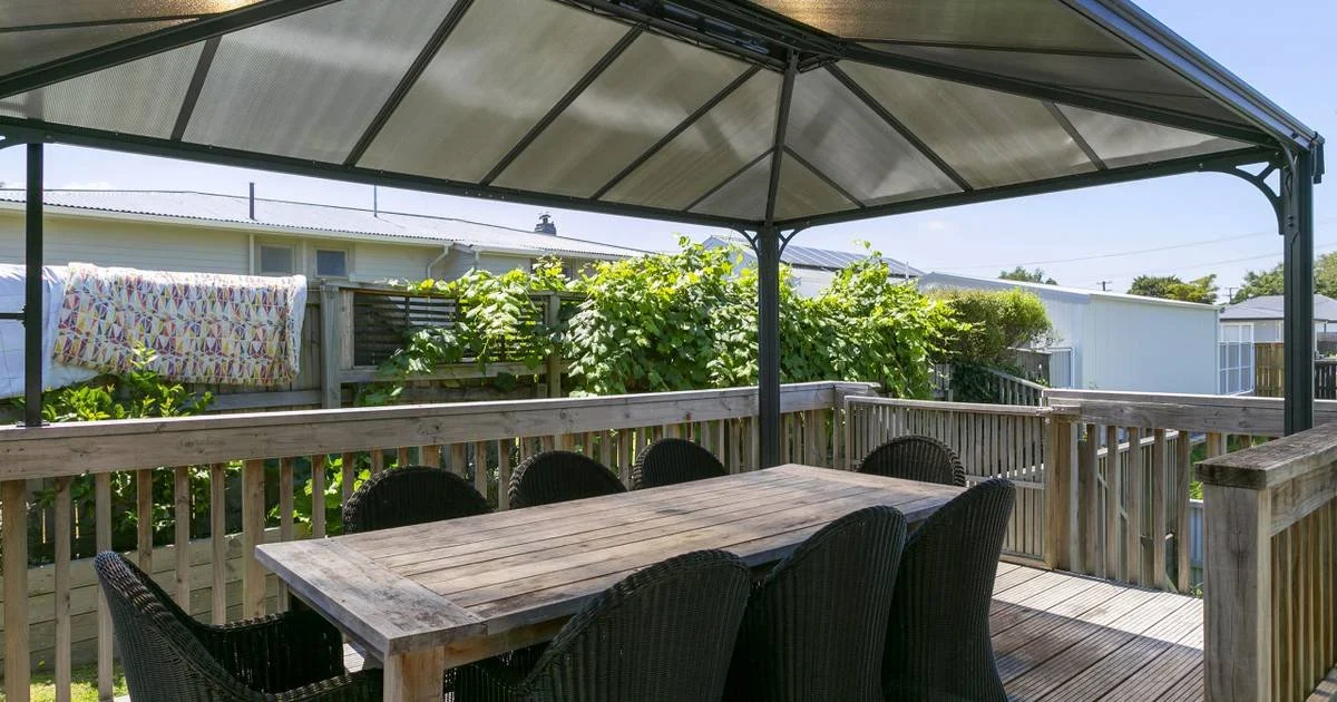 Outdoor wooden deck with a table and six black chairs, covered by a metal gazebo, with a wooden fence, greenery, and neighboring houses in the background.