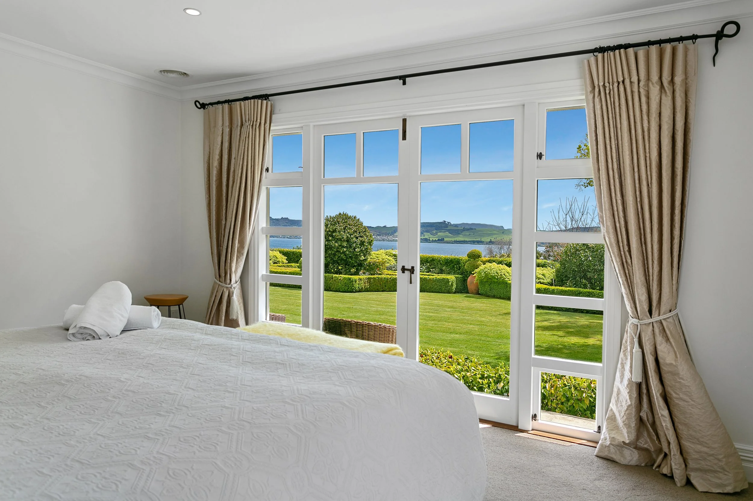A bedroom with large windows and beige curtains, overlooking a green lawn and body of water with hills in the distance.