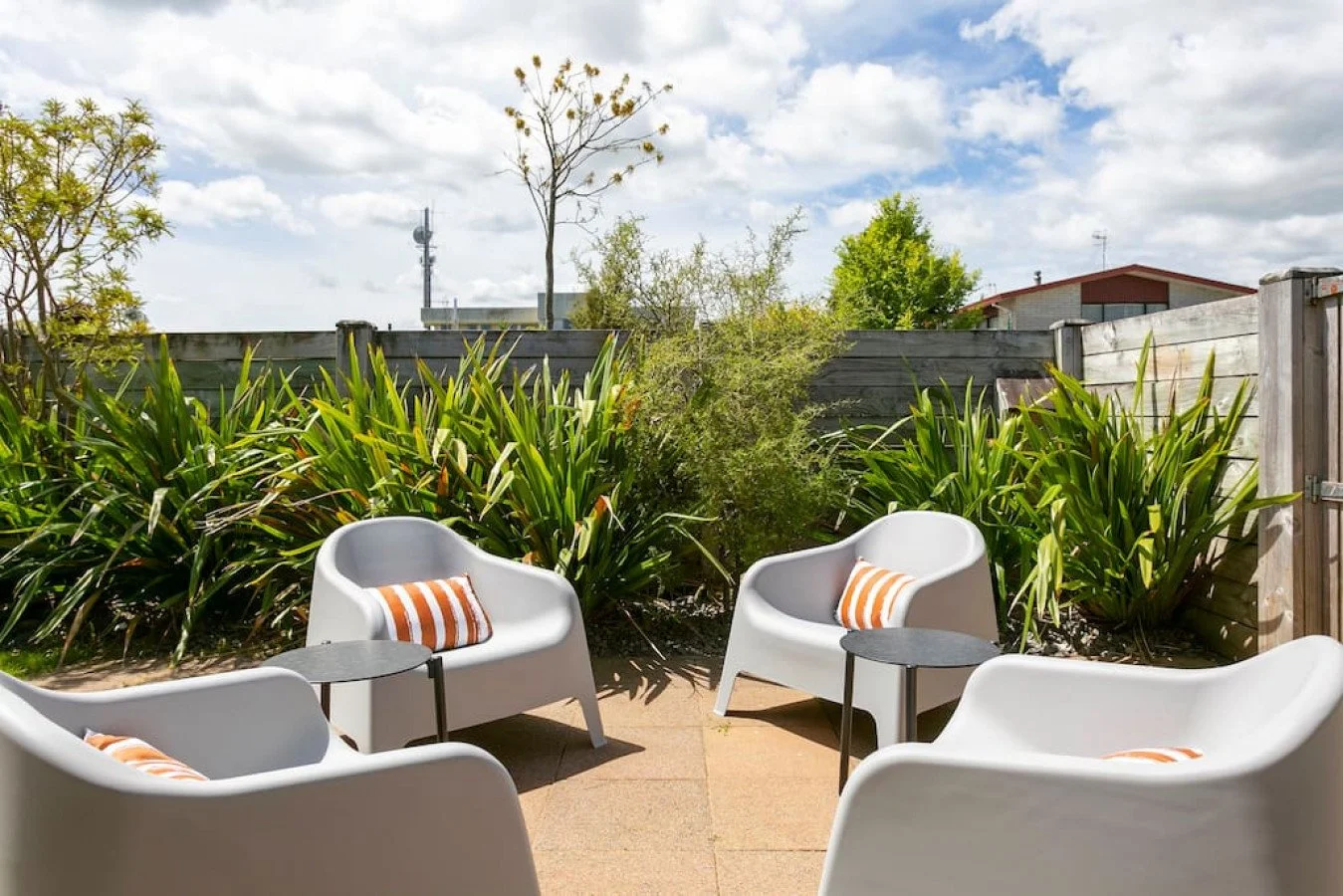 Outdoor patio with four white modern chairs with striped cushions, small side tables, and lush green plants behind a wooden fence under a partly cloudy sky.