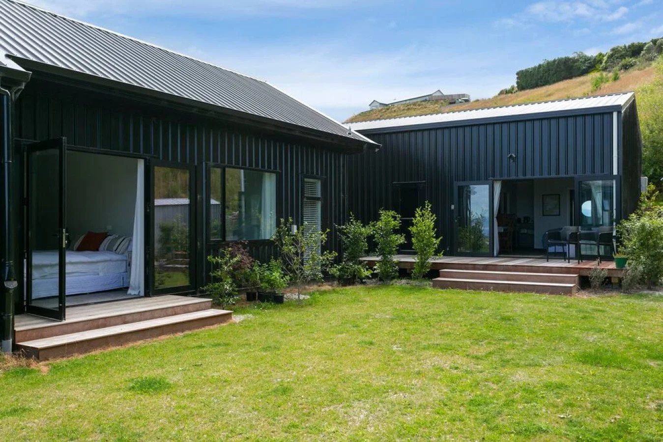 Modern black metal house with large glass doors and a wooden deck, surrounded by green grass and small plants, with a hillside and clear blue sky in the background.