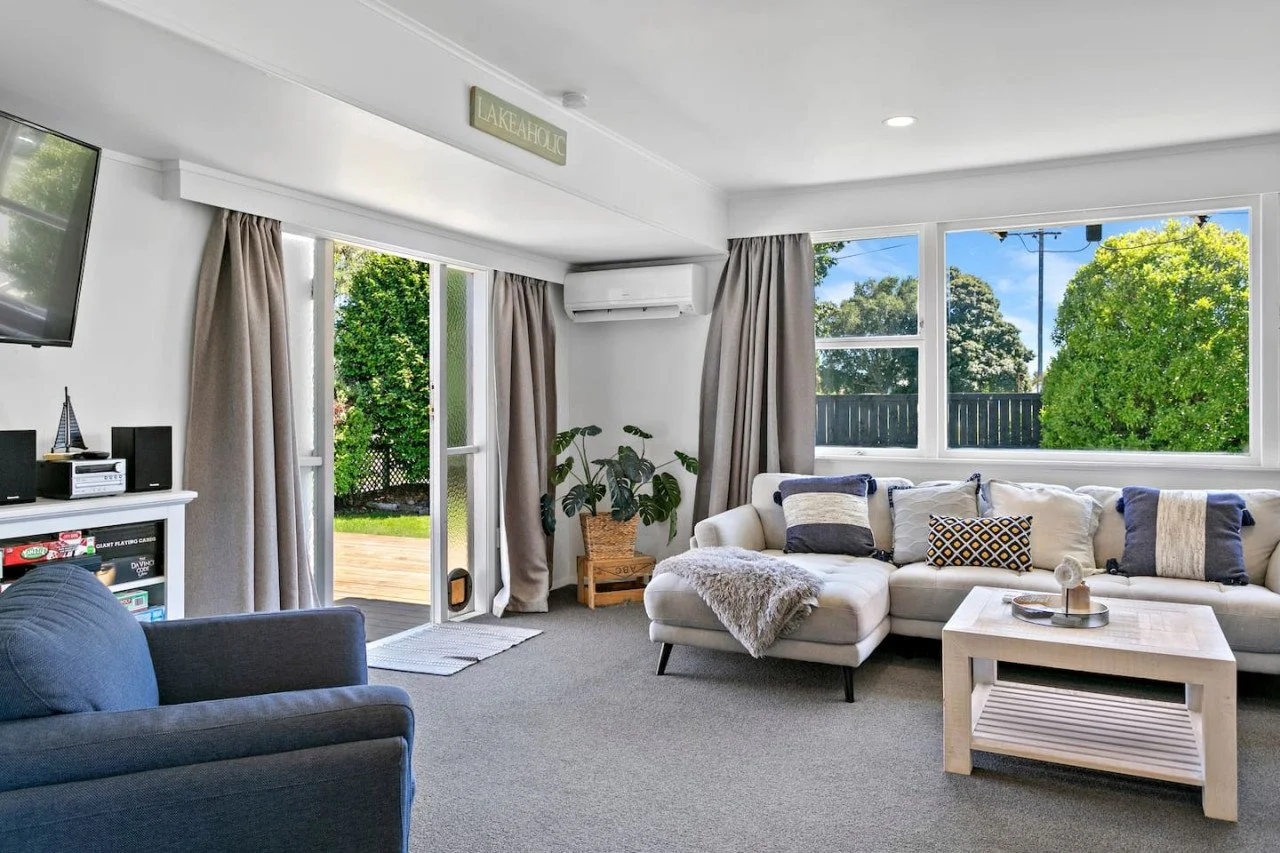 Living room with white sectional sofa, decorative pillows, coffee table, TV mounted on wall, window with curtains, potted plant, and door leading to outdoor deck with trees visible outside.