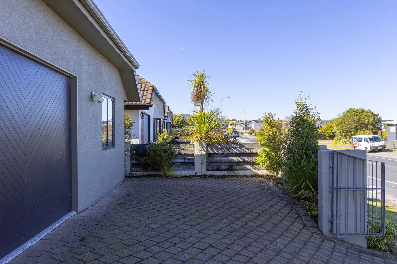 Residential driveway with brick paving, garden plants, a wooden fence, and a gate, next to a gray house, with a street view and a blue sky.
