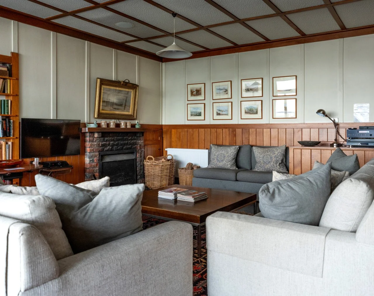 Cozy living room with beige and gray sofas, a wooden coffee table with magazines, a brick fireplace, a mounted TV, framed pictures on the wall, and a bookshelf