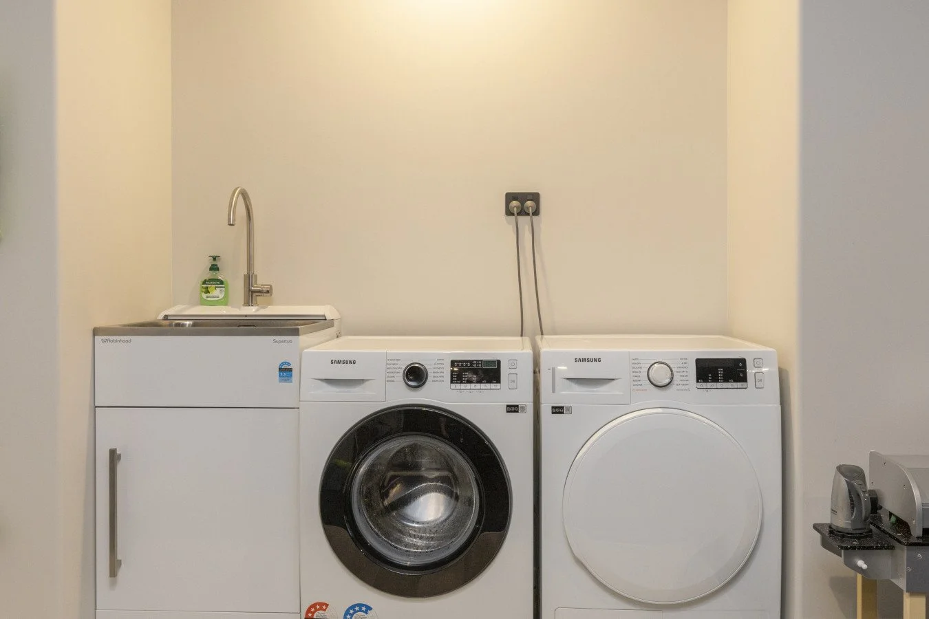Laundry room with a sink, a Samsung washing machine, and a Samsung dryer against a light-colored wall.
