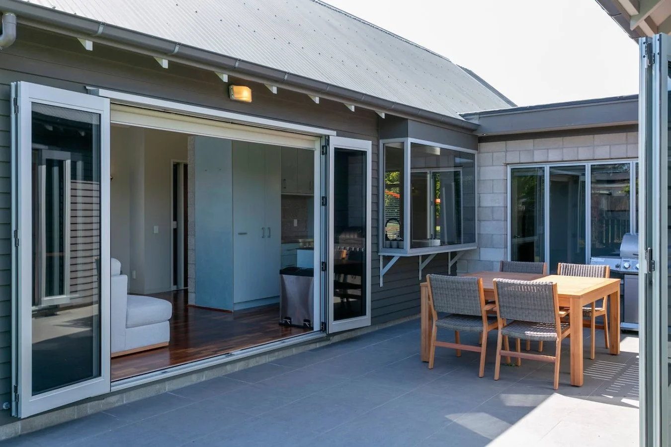 Outdoor patio area with a dining table, six chairs, and a BBQ grill, adjacent to a house with large sliding glass doors.