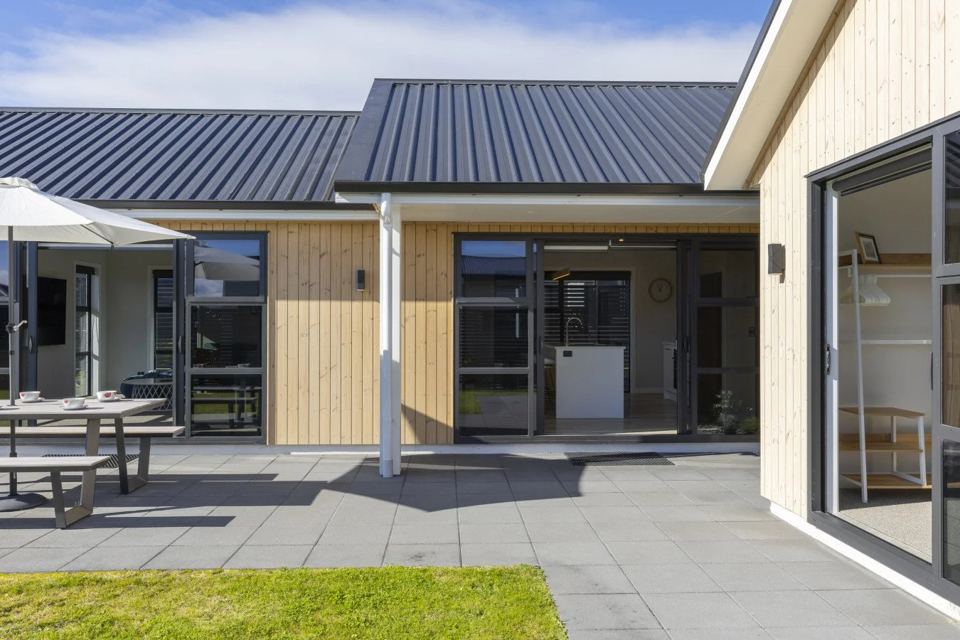 Modern house with light wooden exterior, black metal roof, and large glass doors leading to outdoor patio with furniture and an umbrella.