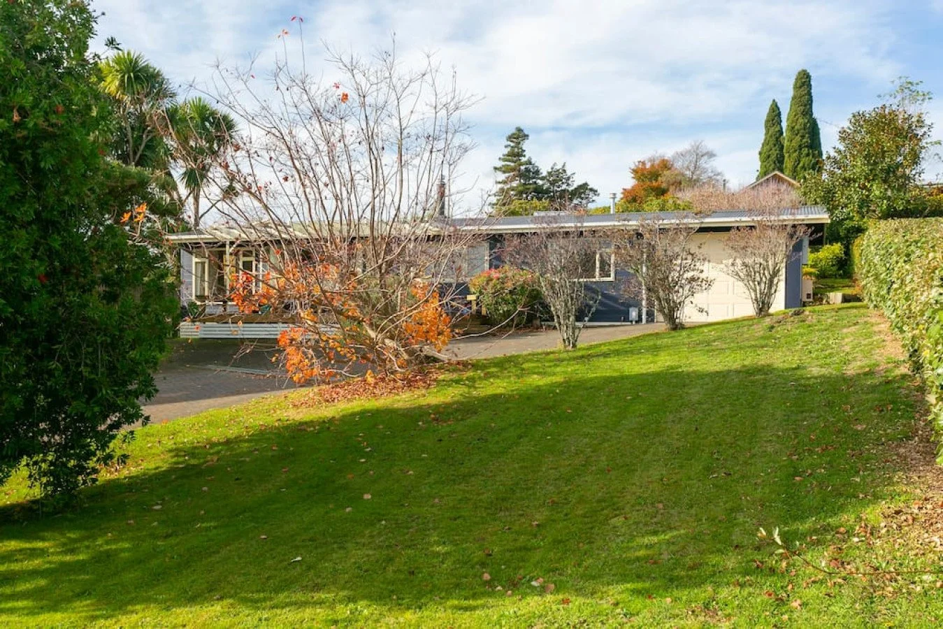 A house with a blue exterior and a white garage door, surrounded by trees and bushes with some leaves fallen, on a sunny day with a partly cloudy sky.
