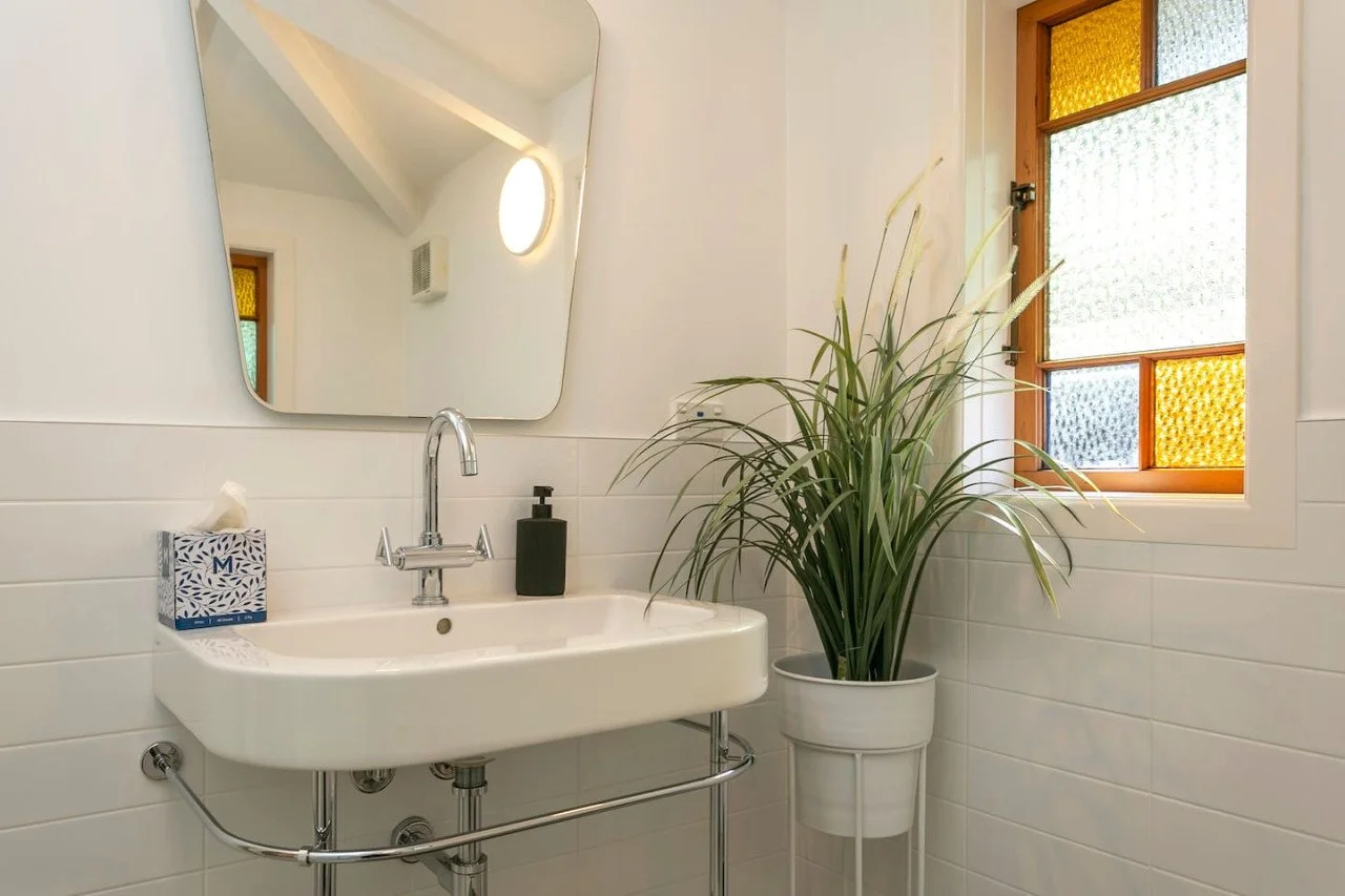 Bathroom with white tile walls, a white sink, a mirror, a black soap dispenser, a tissue box labeled 'M', a green plant in a white pot, and a window with textured glass panes in brown framing.