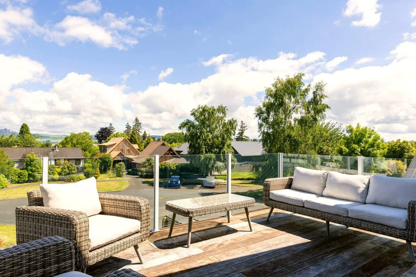 Outdoor patio furniture with wicker sofas and a table on a wooden deck, overlooking a suburban neighborhood with trees and houses under a partly cloudy sky.