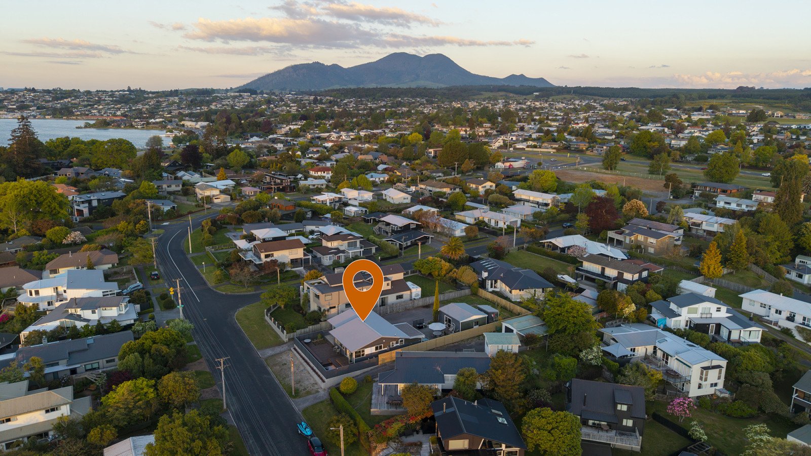 Aerial view of a residential neighborhood with houses, trees, and a lake, with mountains in the background, during sunset.