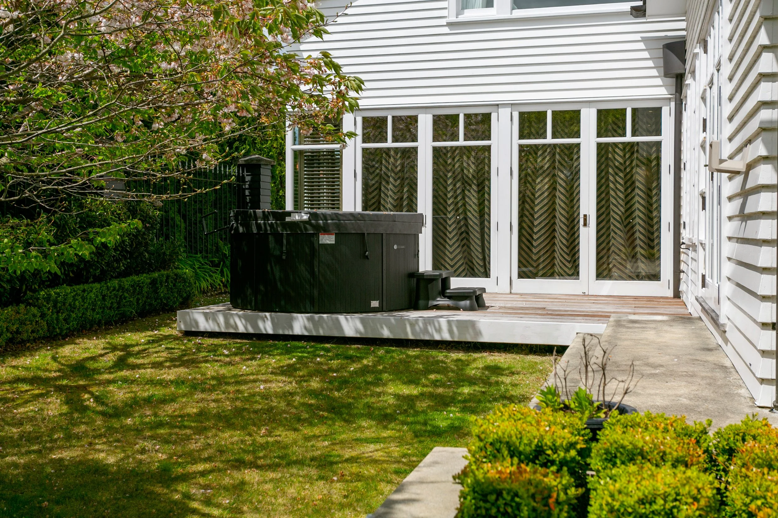 Backyard patio with an above-ground hot tub, wooden deck, sliding glass doors with patterned curtains, and surrounding greenery.
