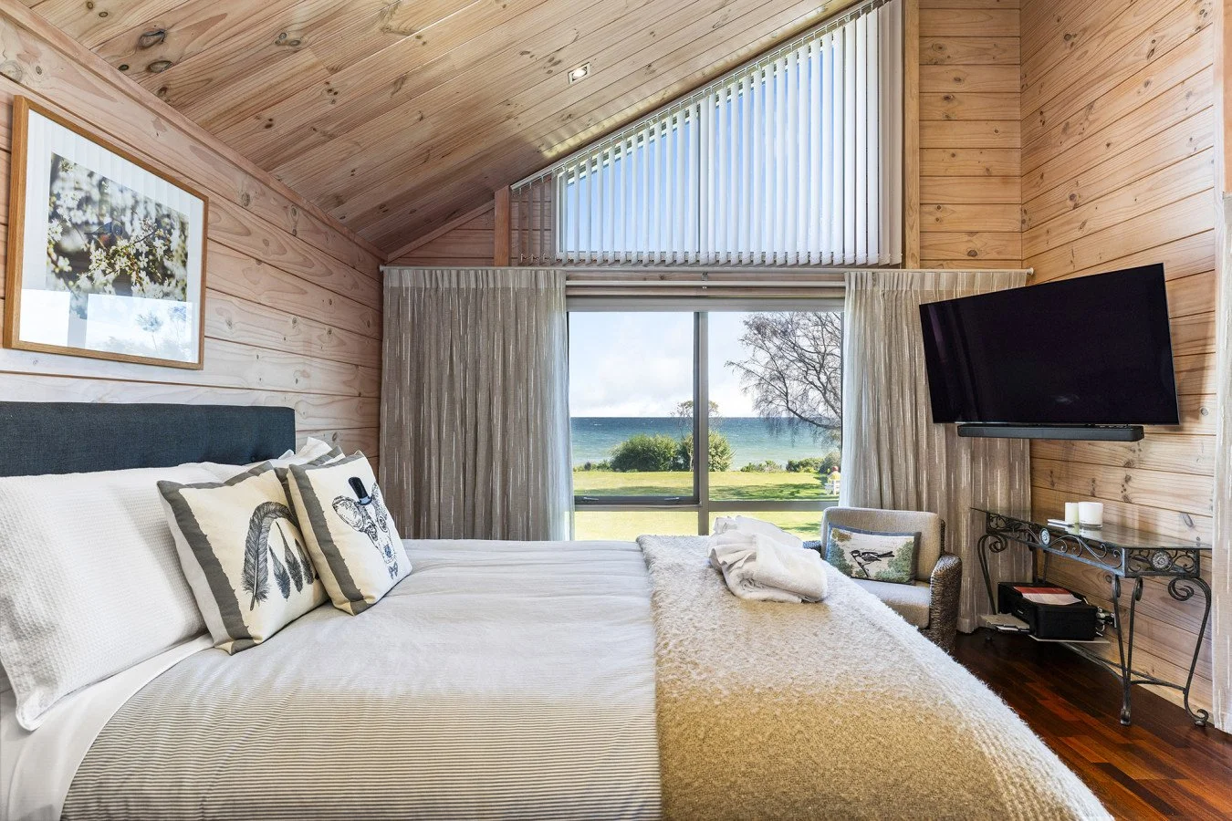 A cozy bedroom with wood-paneled walls and ceiling, featuring a large window with curtains revealing a view of a grassy area, trees, and a body of water. There is a bed with white bedding and decorative pillows, a chair near the window, and a wall-mounted flat-screen TV.