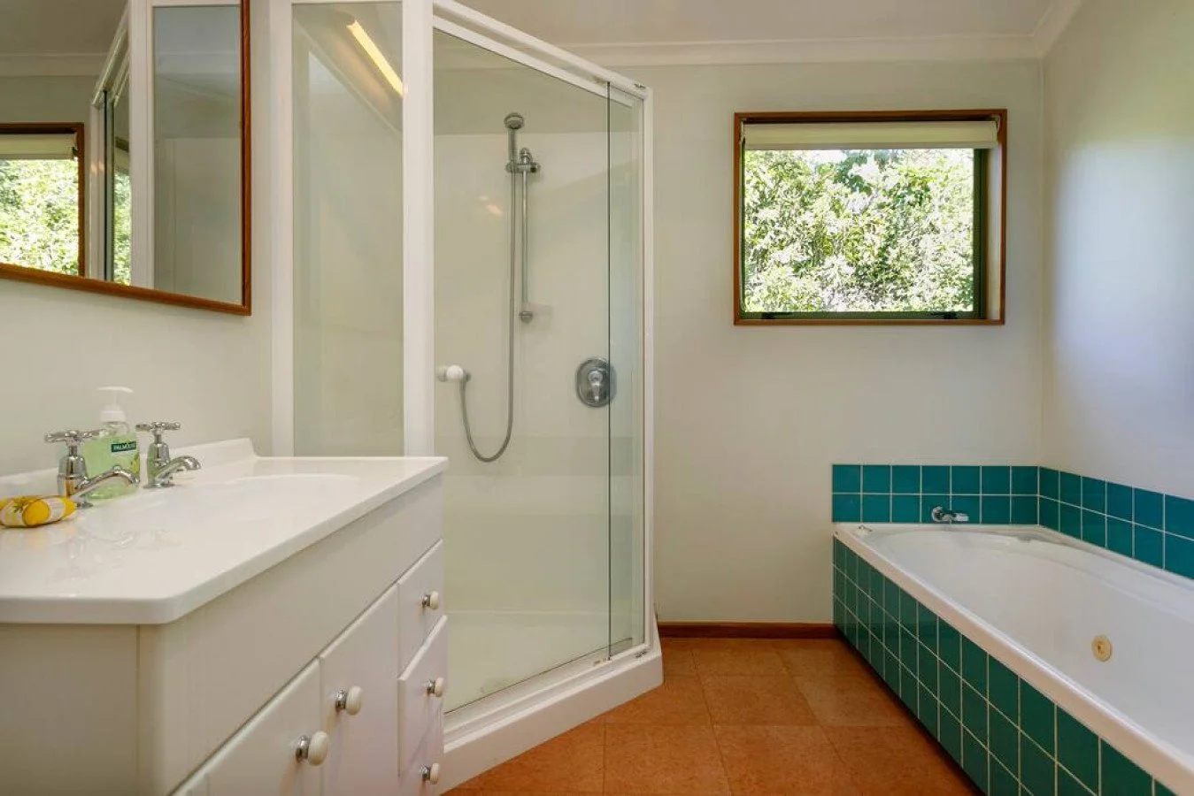 Bathroom with a glass shower stall, a white vanity with a mirror, and a bathtub with teal tiles, and two windows showing greenery outside.