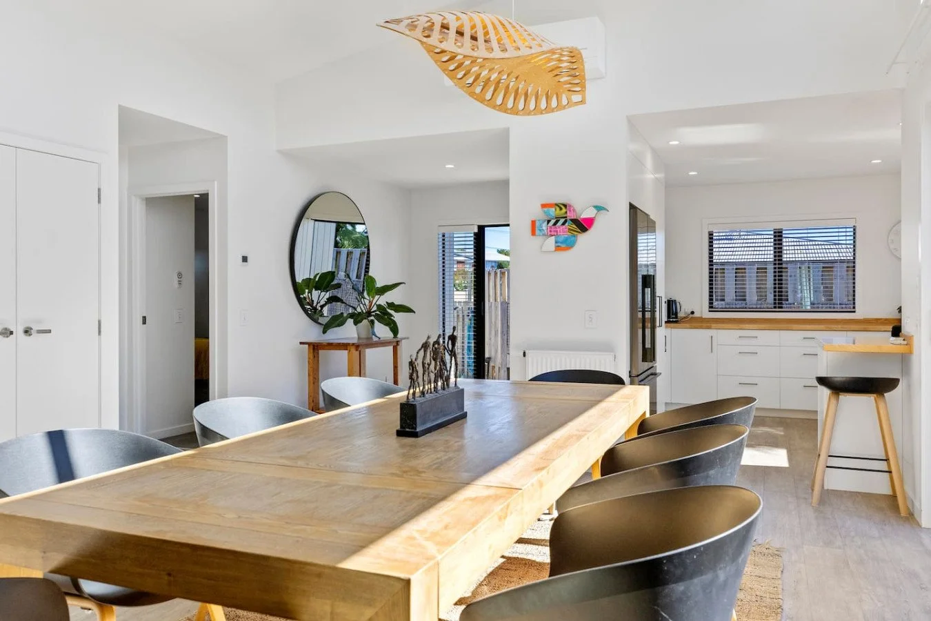 Bright, modern kitchen and dining area with a large wooden table, black and gray chairs, a pendant light, a wall mirror, colorful wall art, and view of the outside through windows and a door.