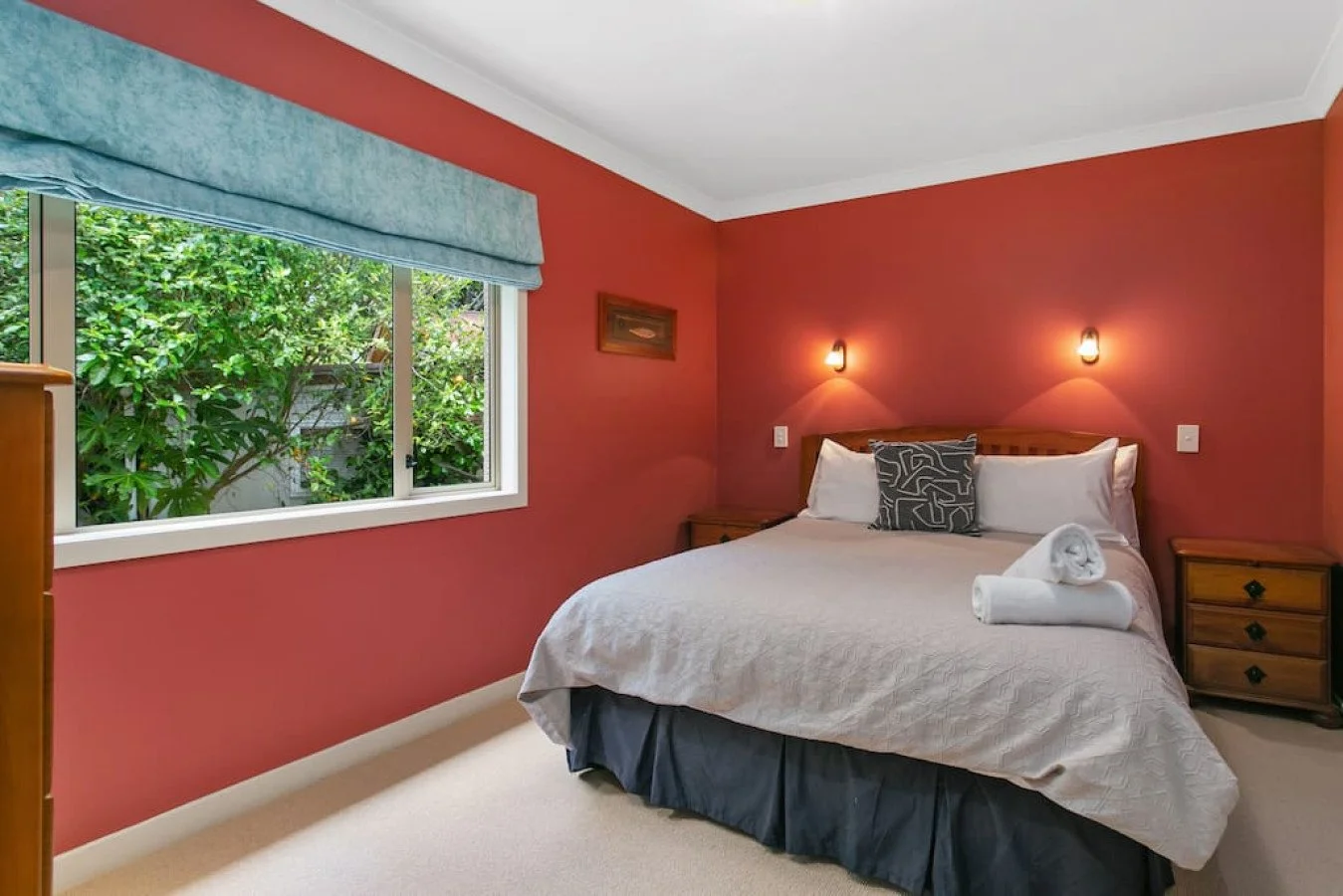 A cozy bedroom with a bed against a red wall, two wall-mounted lights, a window with greenery outside, and furniture including a nightstand and a dresser.