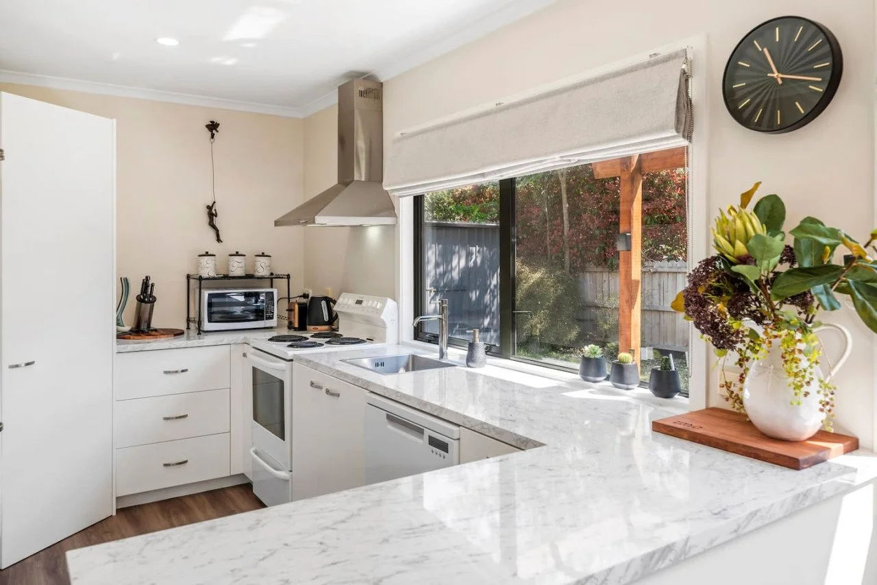 Bright kitchen with white cabinets and marble countertops, a window with a view of a backyard with trees, a black wall clock, a potted plant on the counter, and kitchen appliances including a microwave, oven, and stovetop.