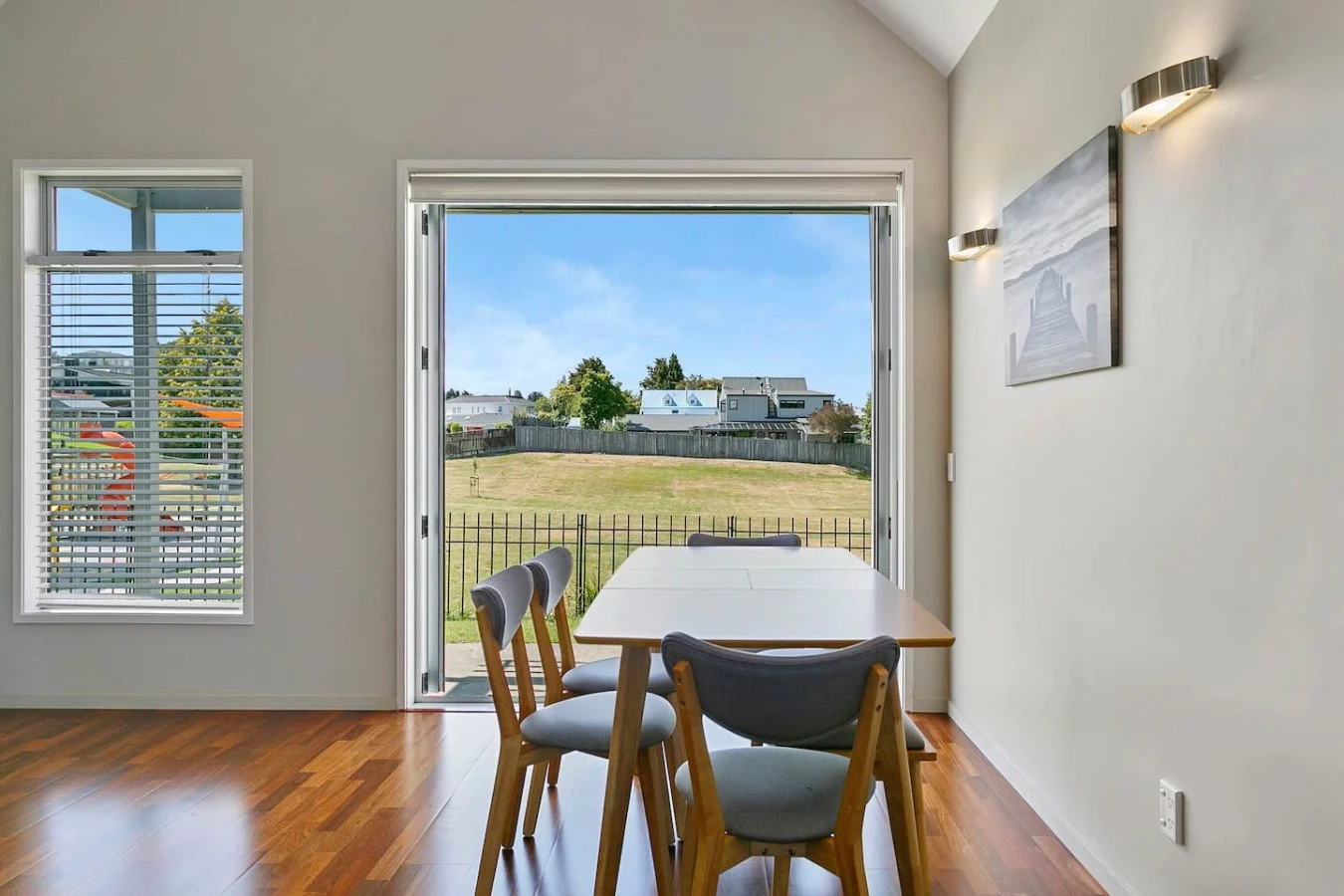 Dining area with a wooden table and four chairs, open to a backyard with a grassy yard, a black fence, and houses in the distance under a blue sky.