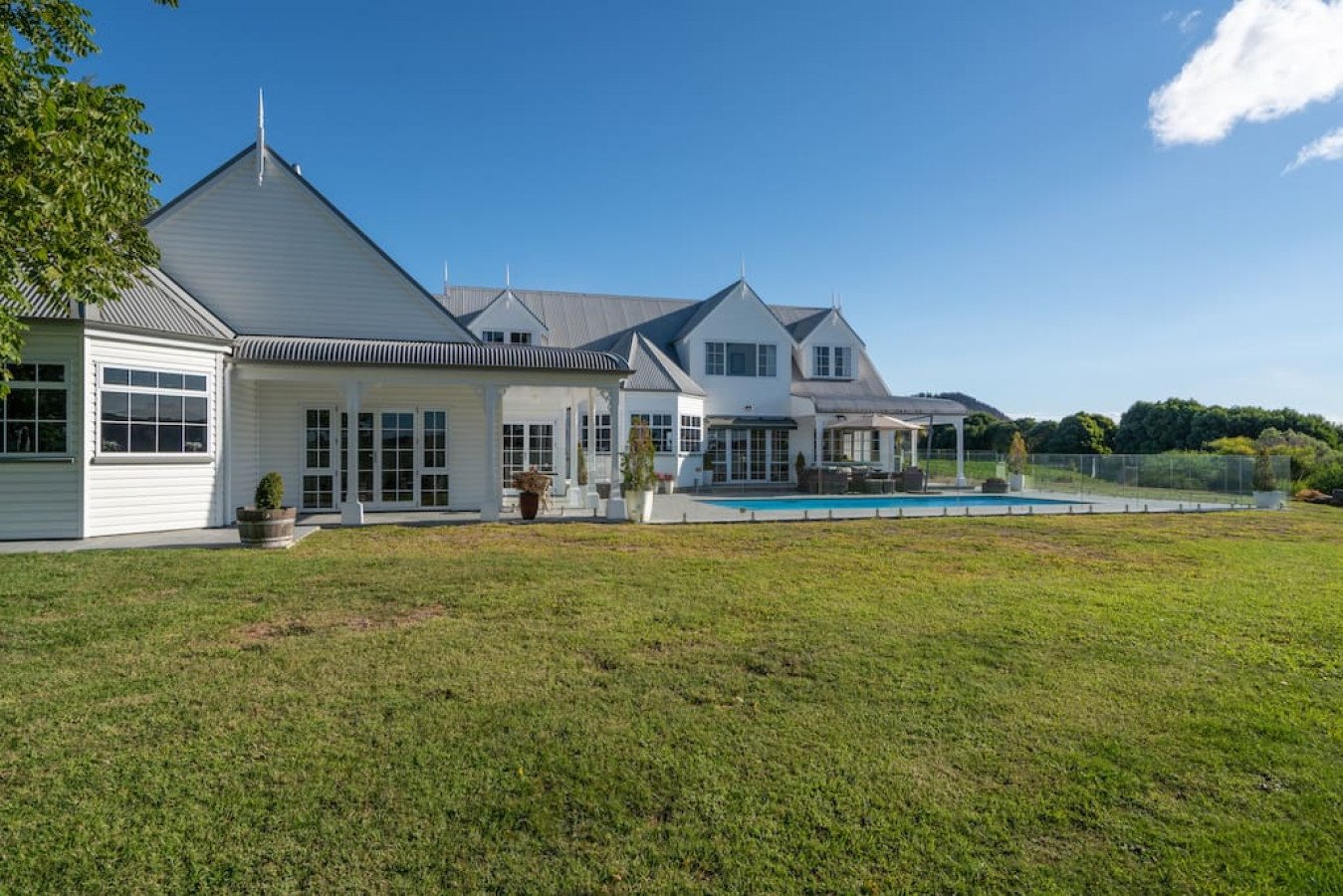 Large white house with multiple gabled roofs and a swimming pool in the backyard, green lawn, and clear blue sky.