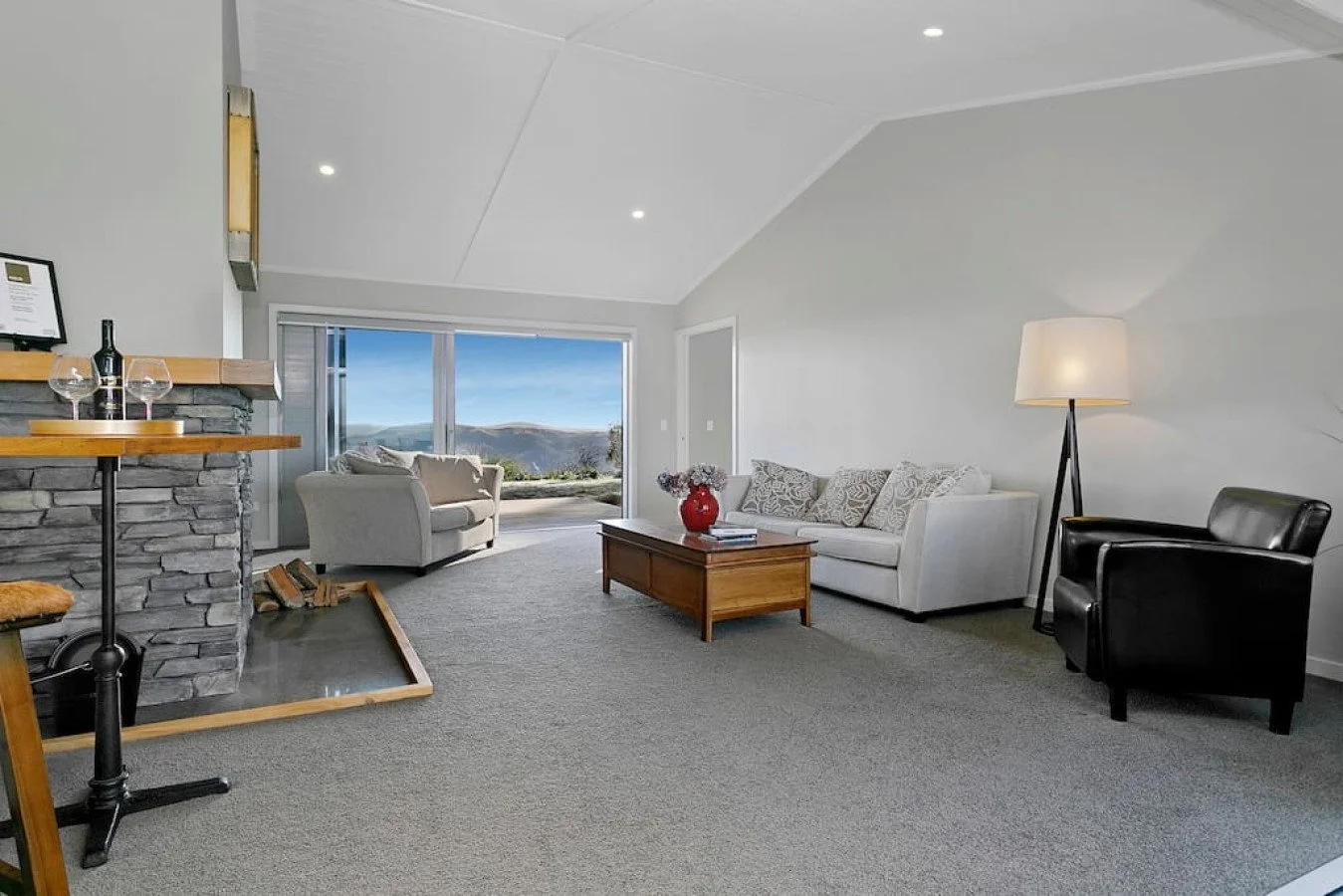 A spacious living room with white walls and ceiling, grey carpet, a white sofa with patterned cushions, a black armchair, a wooden coffee table with a red vase, an off-white armchair near large glass sliding doors, and landscape view of hills outside