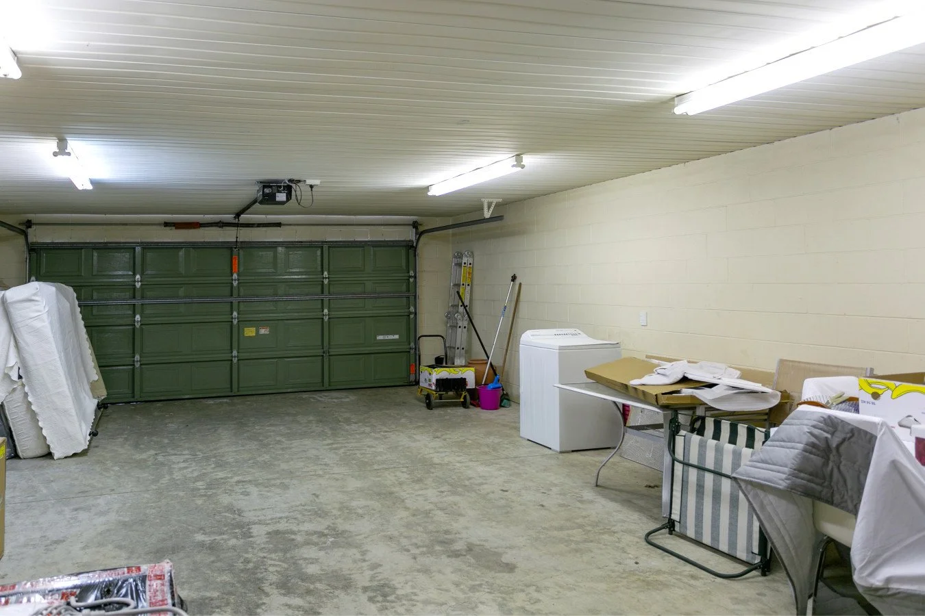 Empty garage with a green garage door, cleaning supplies, and furniture covered with sheets.
