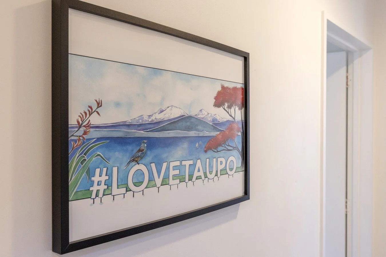 Framed watercolor painting on a white wall featuring a mountain landscape, blue lake, red trees, and the hashtag #LOVETAUPO.