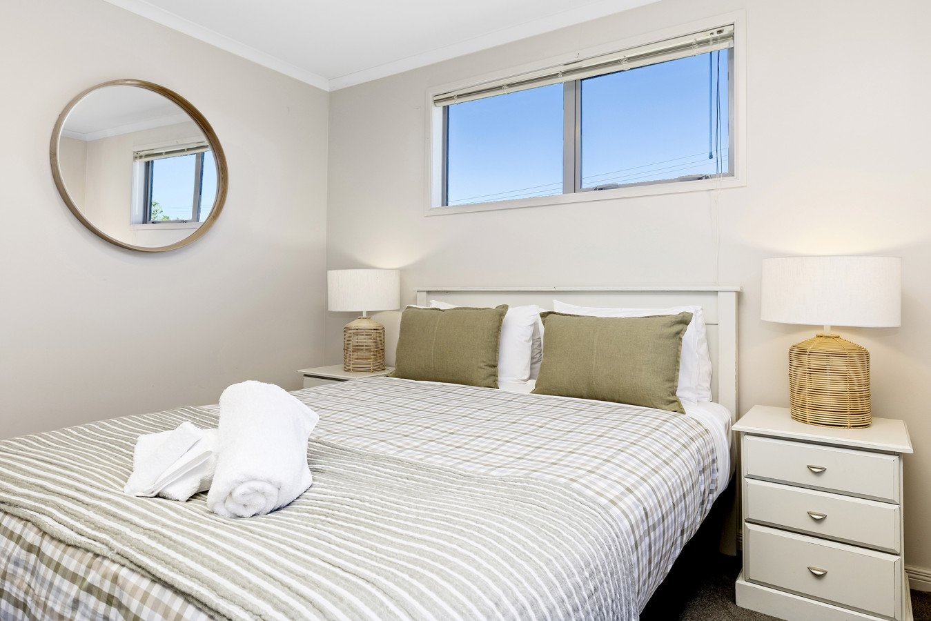A neatly made bed with a striped beige and white bedspread, two green pillows, and two white pillows, in a bright bedroom with white walls, a window, and two bedside tables with wicker lamps. Towels are rolled and placed on the bed.