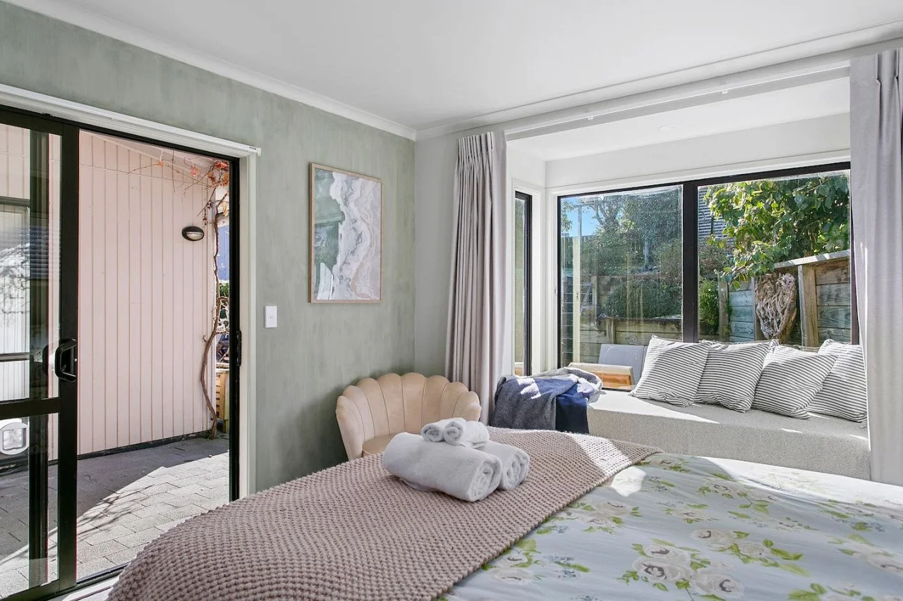 A bright bedroom with large windows letting in natural light, a bed with floral sheets and rolled towels on top, a white built-in window seat with striped pillows, a beige armchair, and outdoor patio visible through the sliding glass door.
