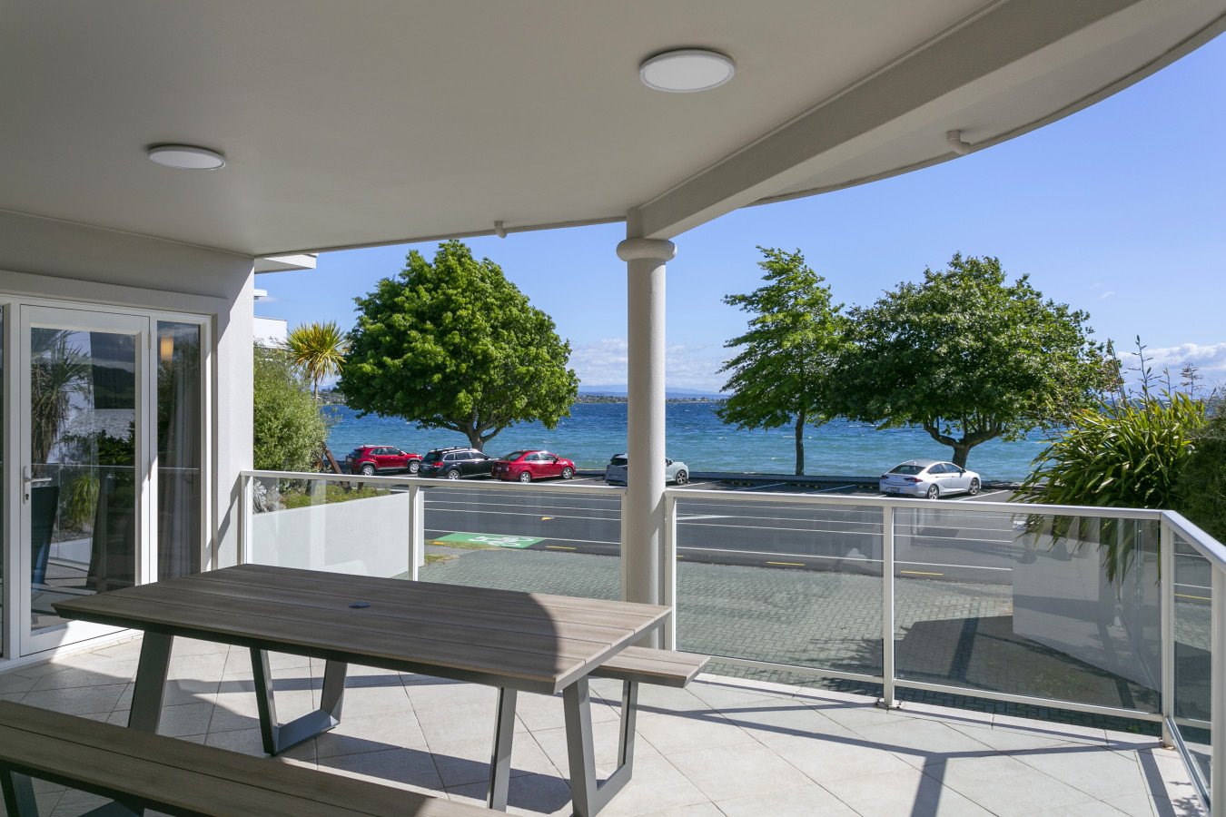 Balcony view overlooking a street, trees, and water with a clear blue sky in the background. There is a wooden table and a bench on the balcony.