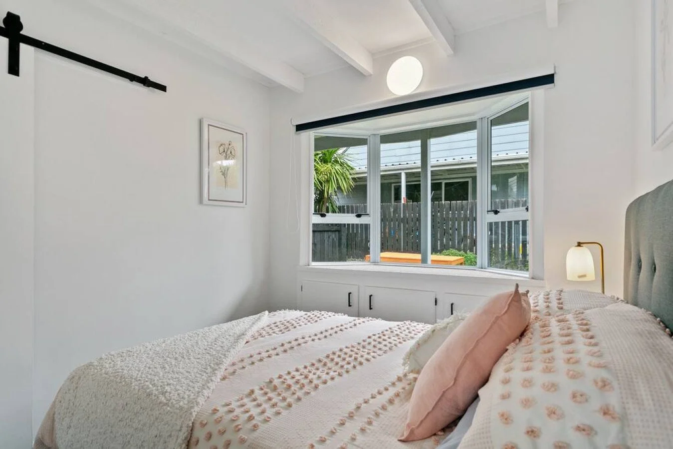 Bedroom with a large window overlooking a backyard with a wooden privacy fence and trees, a bed with pink and white bedding and a pink pillow, a wall-mounted lamp, and a framed art piece of a bicycle.