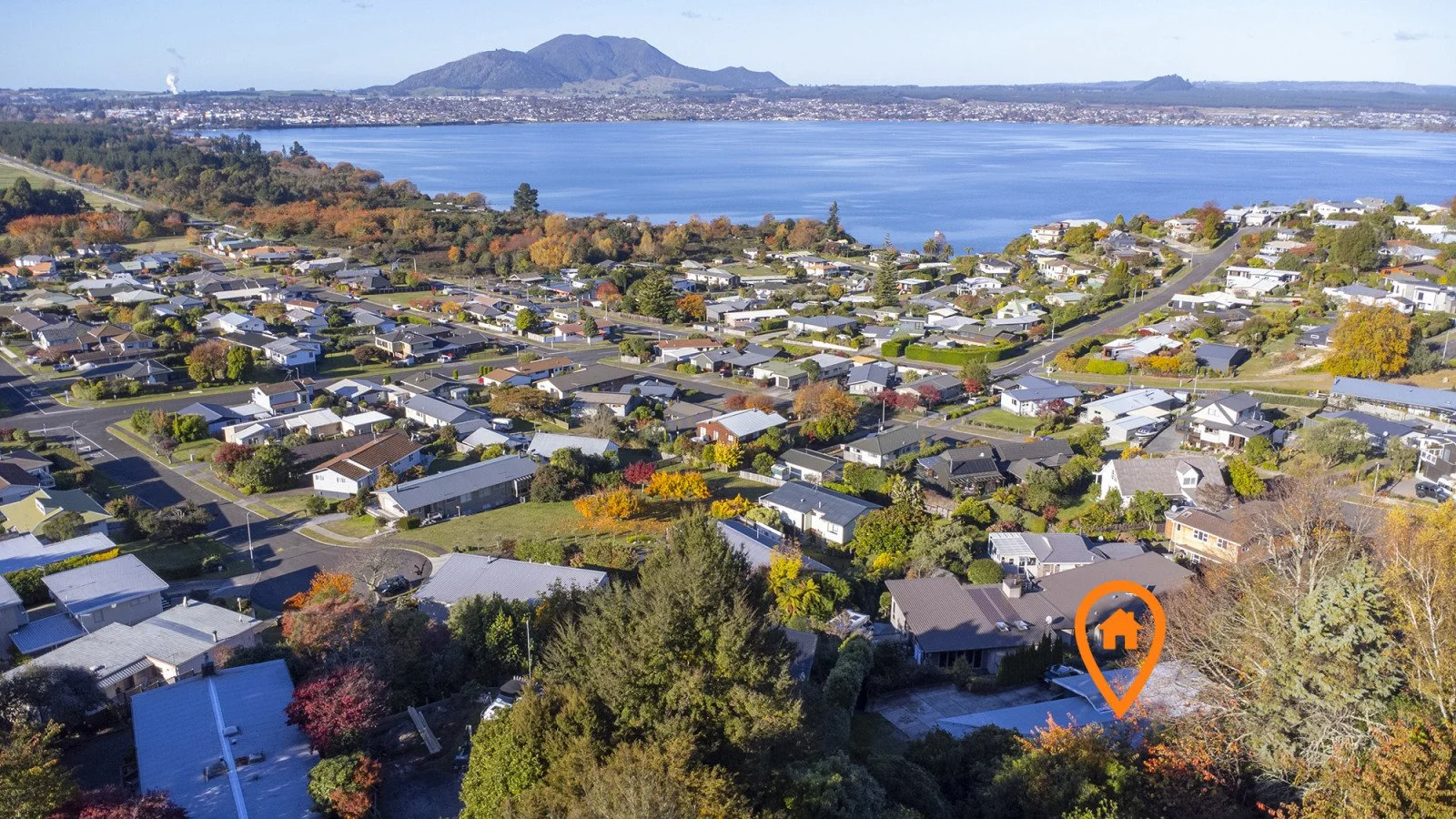 Aerial view of a lakeside residential neighborhood with houses, trees, and a large body of water, with mountains in the background.