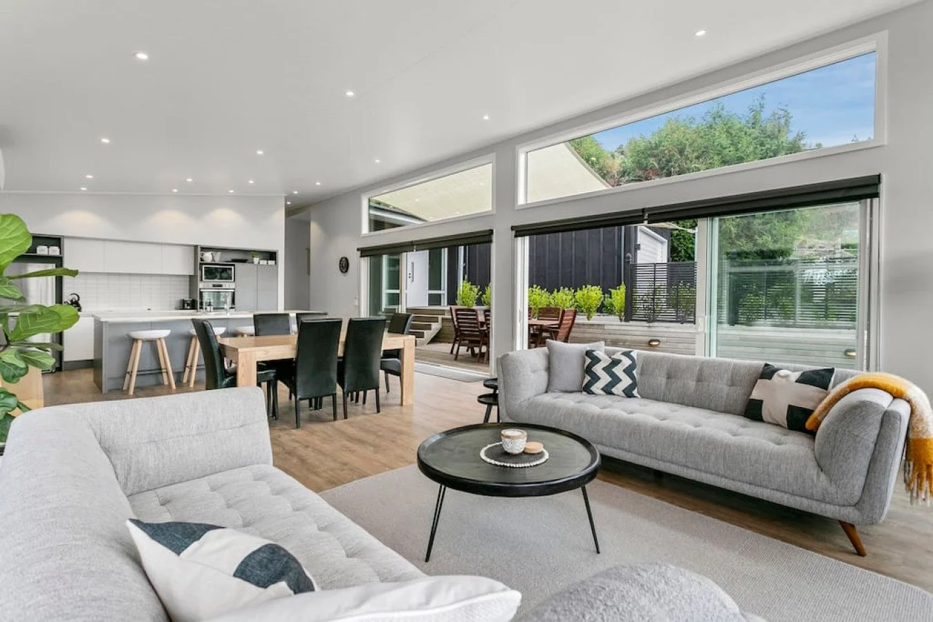 Spacious open-concept living room with large windows, gray sofas, a black coffee table, and a view of the outdoor patio with wooden furniture and greenery.