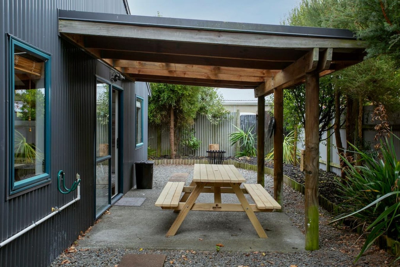 Backyard patio with a wooden picnic table under a covered area, surrounded by greenery and a fence.