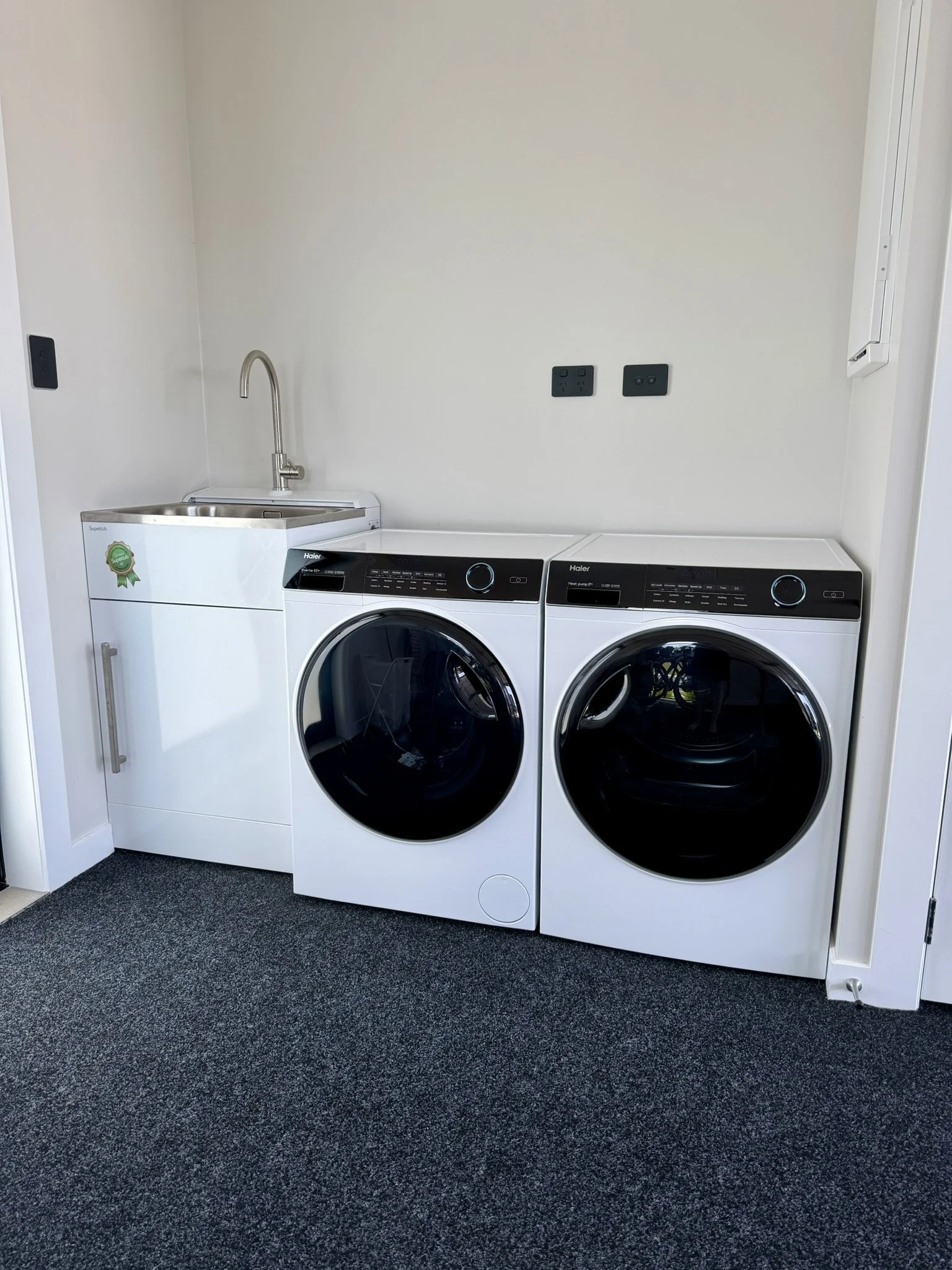 Laundry room with white washer and dryer side by side, small white sink with a silver faucet on the left, and two black electrical outlets on the wall.