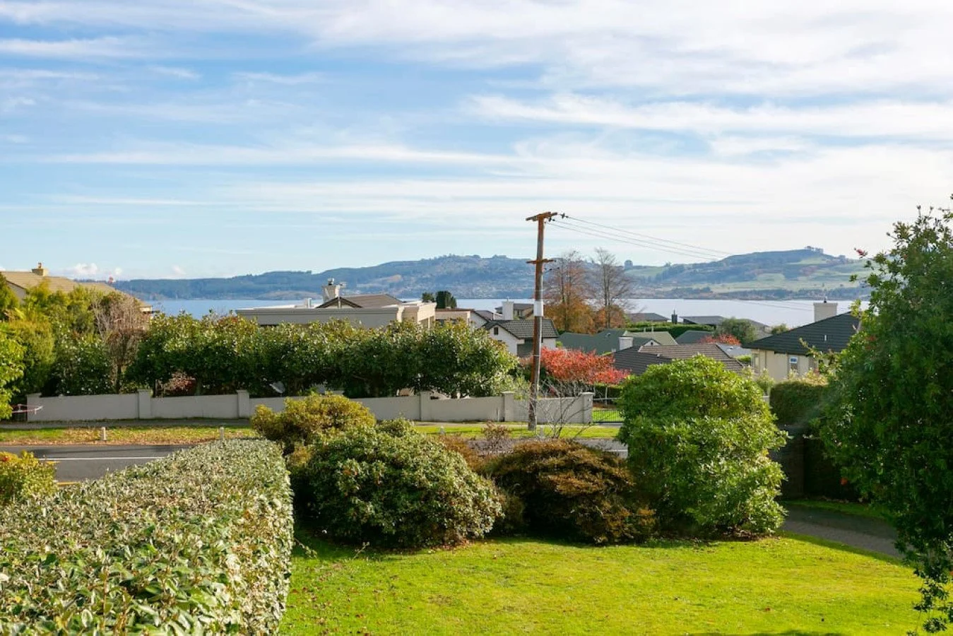 Residential neighborhood with greenery, bushes, trees, and houses, overlooking a body of water with hilly landscape in the background.