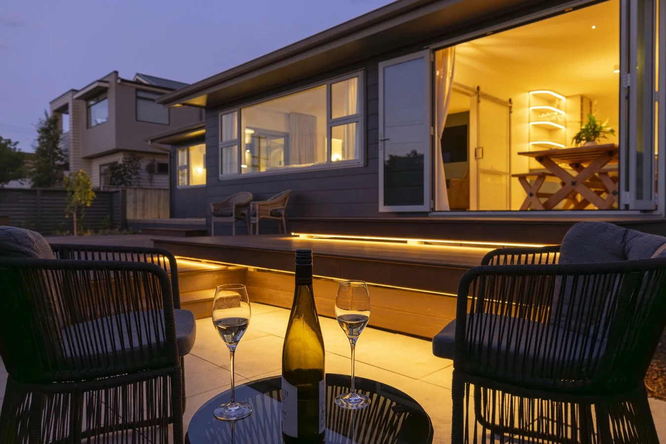 Backyard patio at dusk with two black chairs, a small round table with a wine bottle and two glasses, and a modern house with lit interior and large windows.