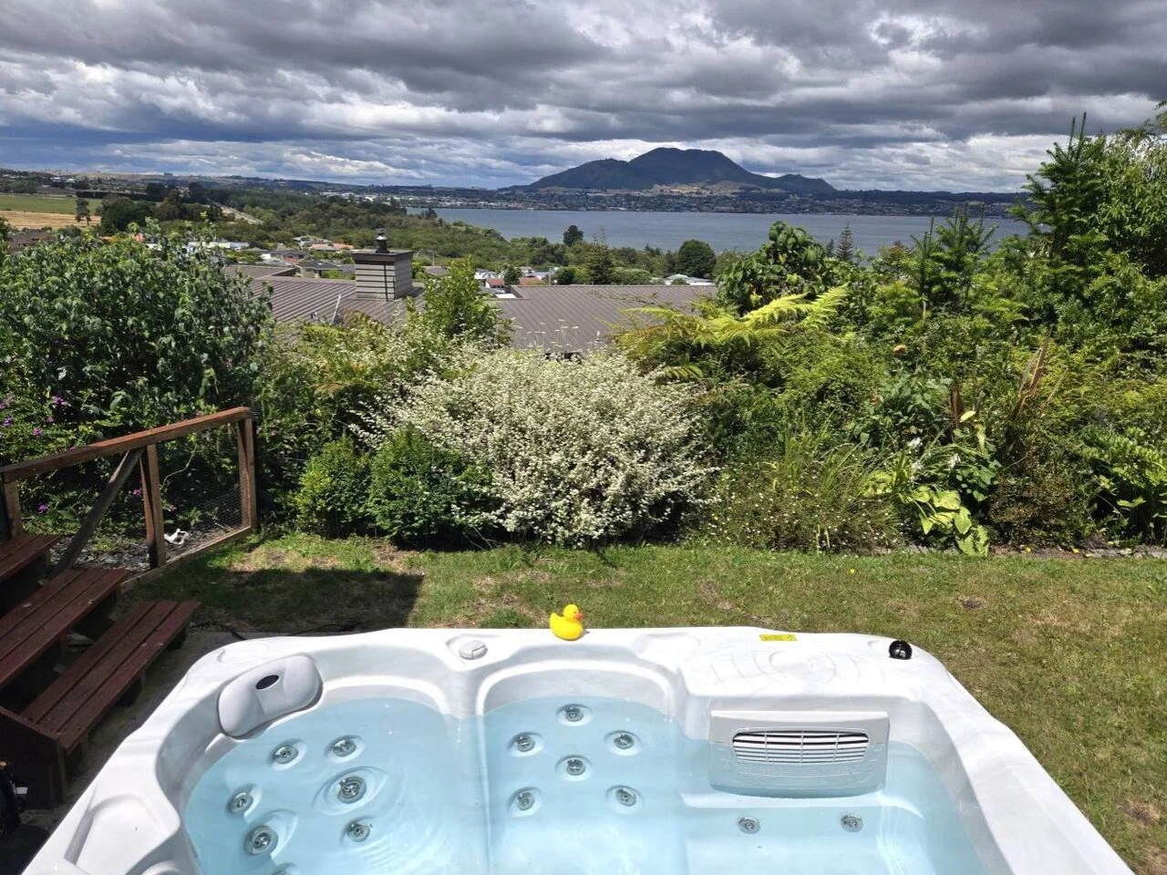 A hot tub with water and jets, a rubber duck, and a small black object on its edge, overlooking a lush green garden with shrubs and trees, with a large lake and mountain in the background under a partly cloudy sky.