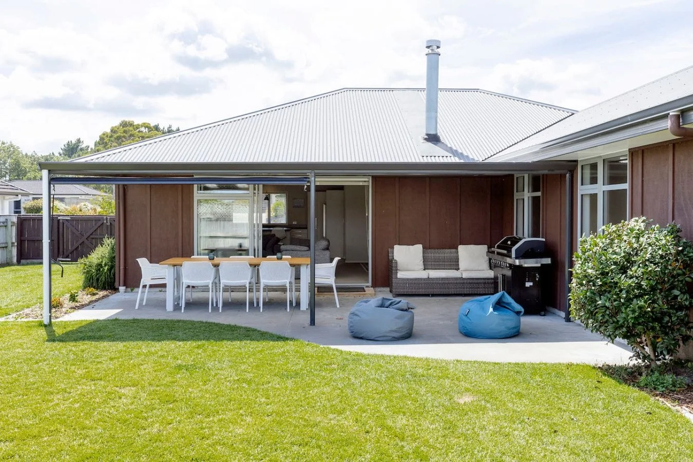 Backyard patio with outdoor dining table, chairs, a sofa, and a barbecue grill on a concrete slab, with a house and green lawn in the background.