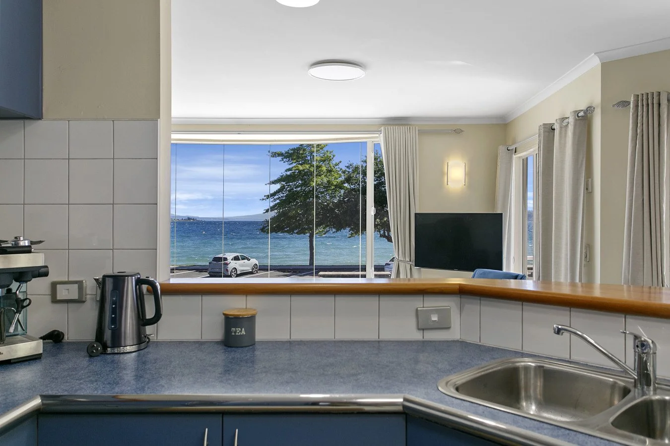 View of a coastal scene through a kitchen window, featuring a car parked near the beach, a tree, and water with a clear sky.
