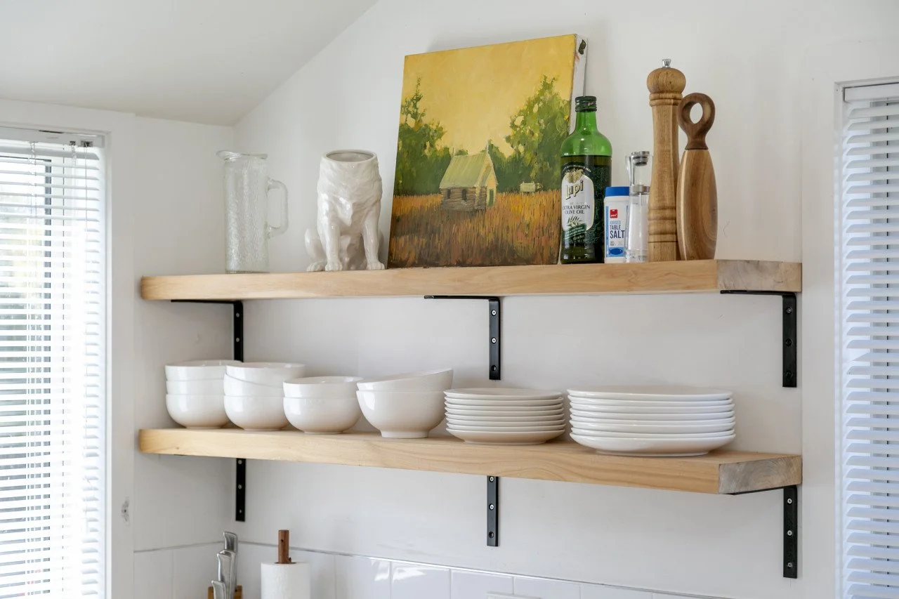 Open kitchen shelves displaying bowls, plates, a painting, olive oil, salt, and utensils, with blinds on windows to the sides.